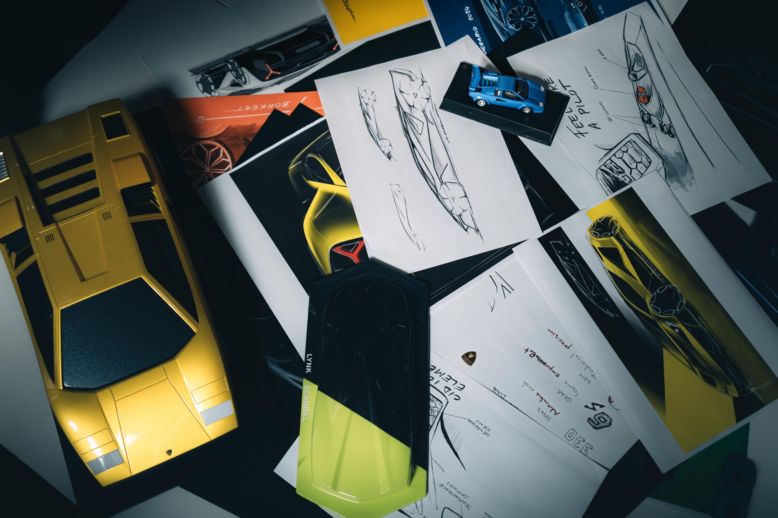 Lamborghini design sketches on a table alongside a yellow Countach scale model and a small blue concept model