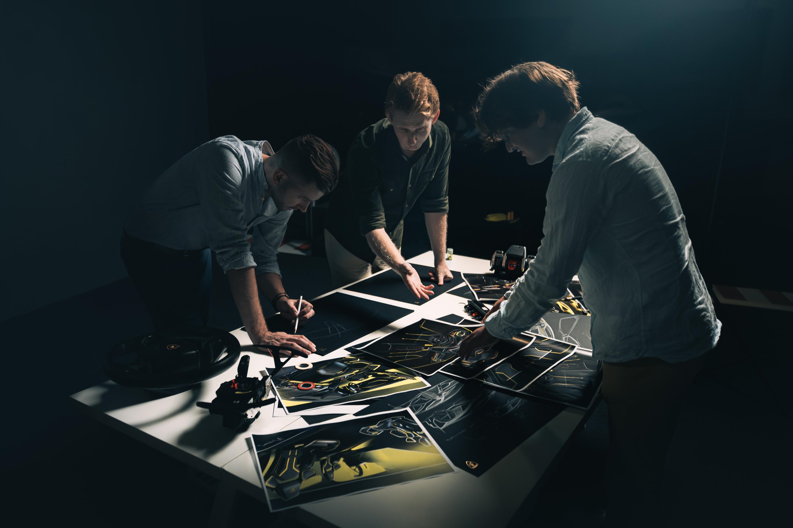 Three Lamborghini designers collaborating on sketches and a concept model in a dimly lit studio