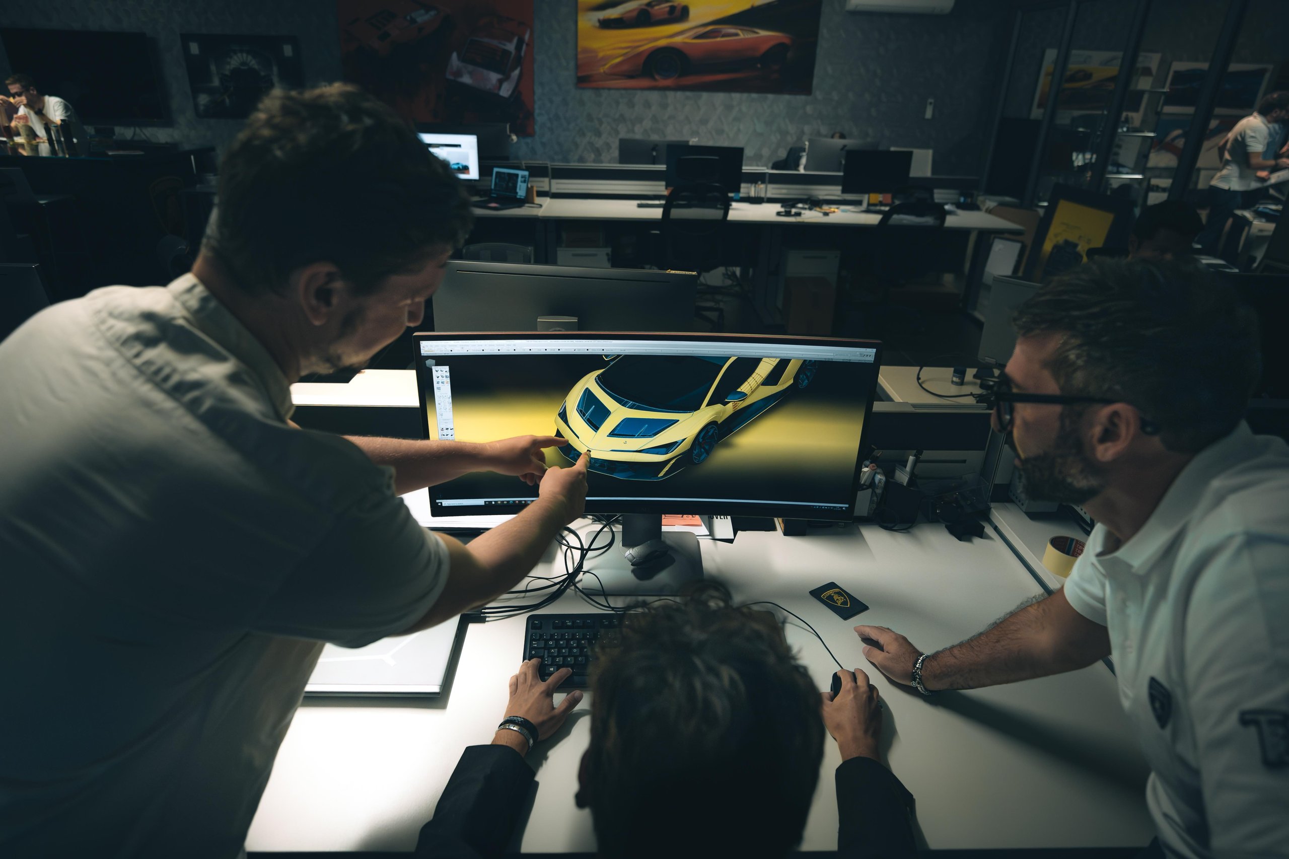 Three Lamborghini designers collaborating around a computer screen displaying a 3D render of a yellow concept car