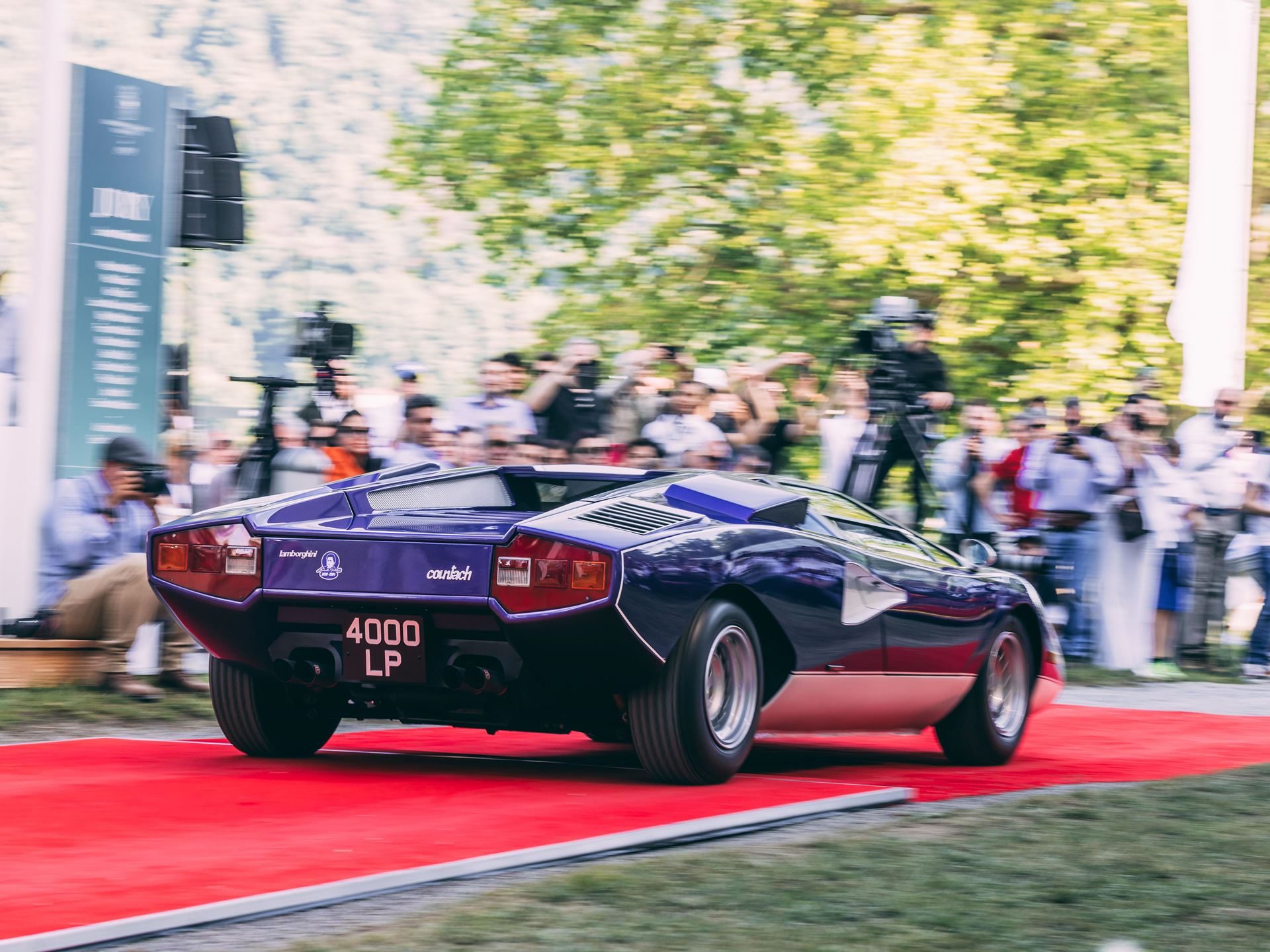Blue lamborghini countach lp400 driving on a red carpet at an outdoor concours event