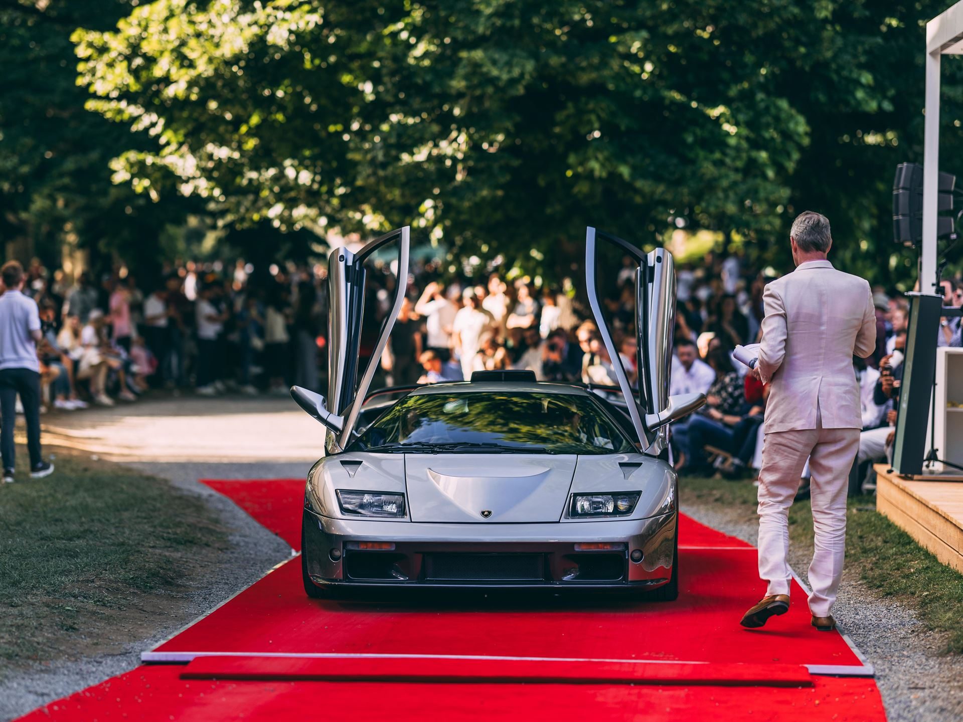 Silver lamborghini diablo gt with scissor doors open on a red carpet at a concours event, with spectators in the background