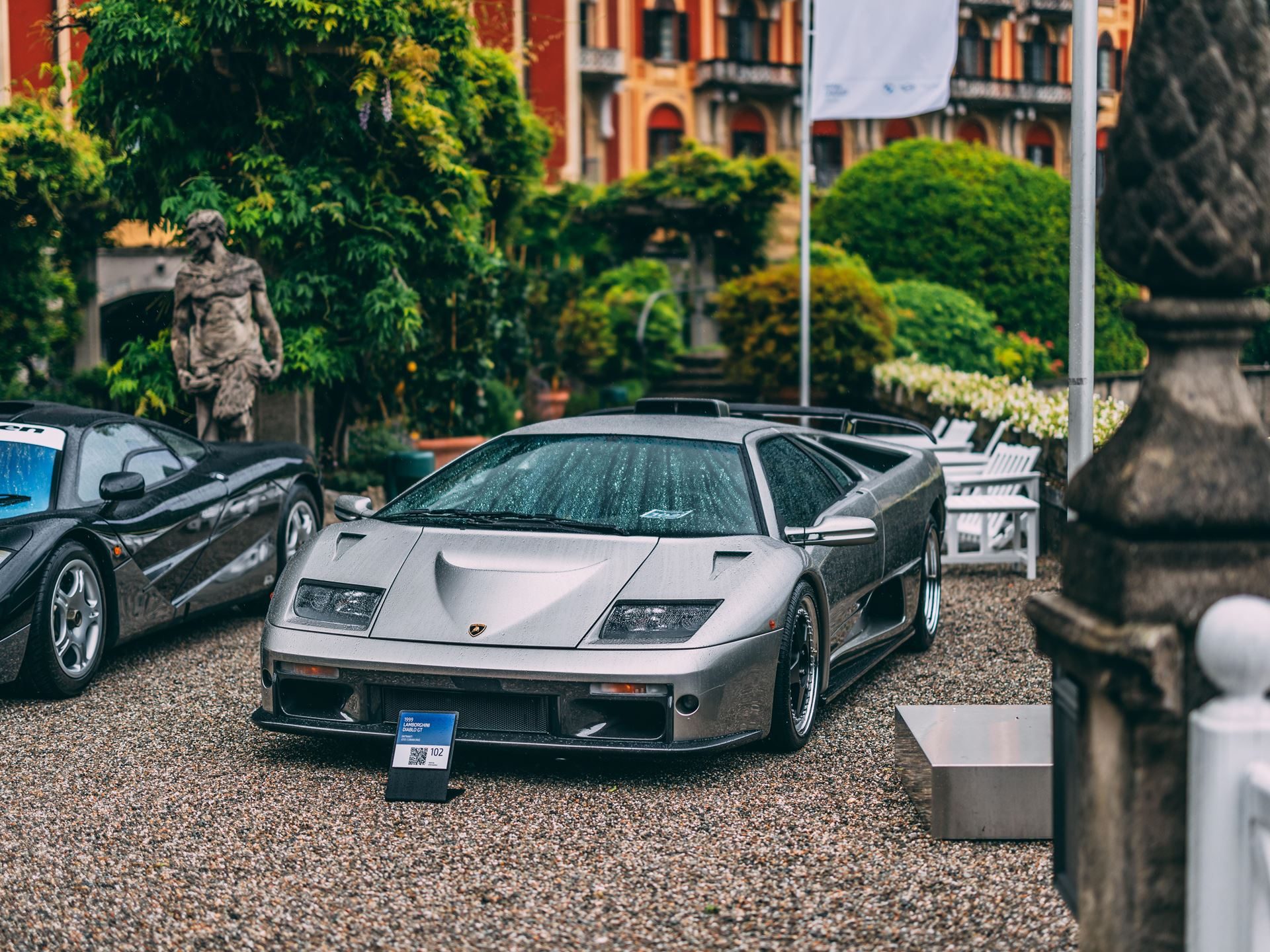 Silver lamborghini diablo parked on gravel at an outdoor concours event with a mclaren f1 visible in the background