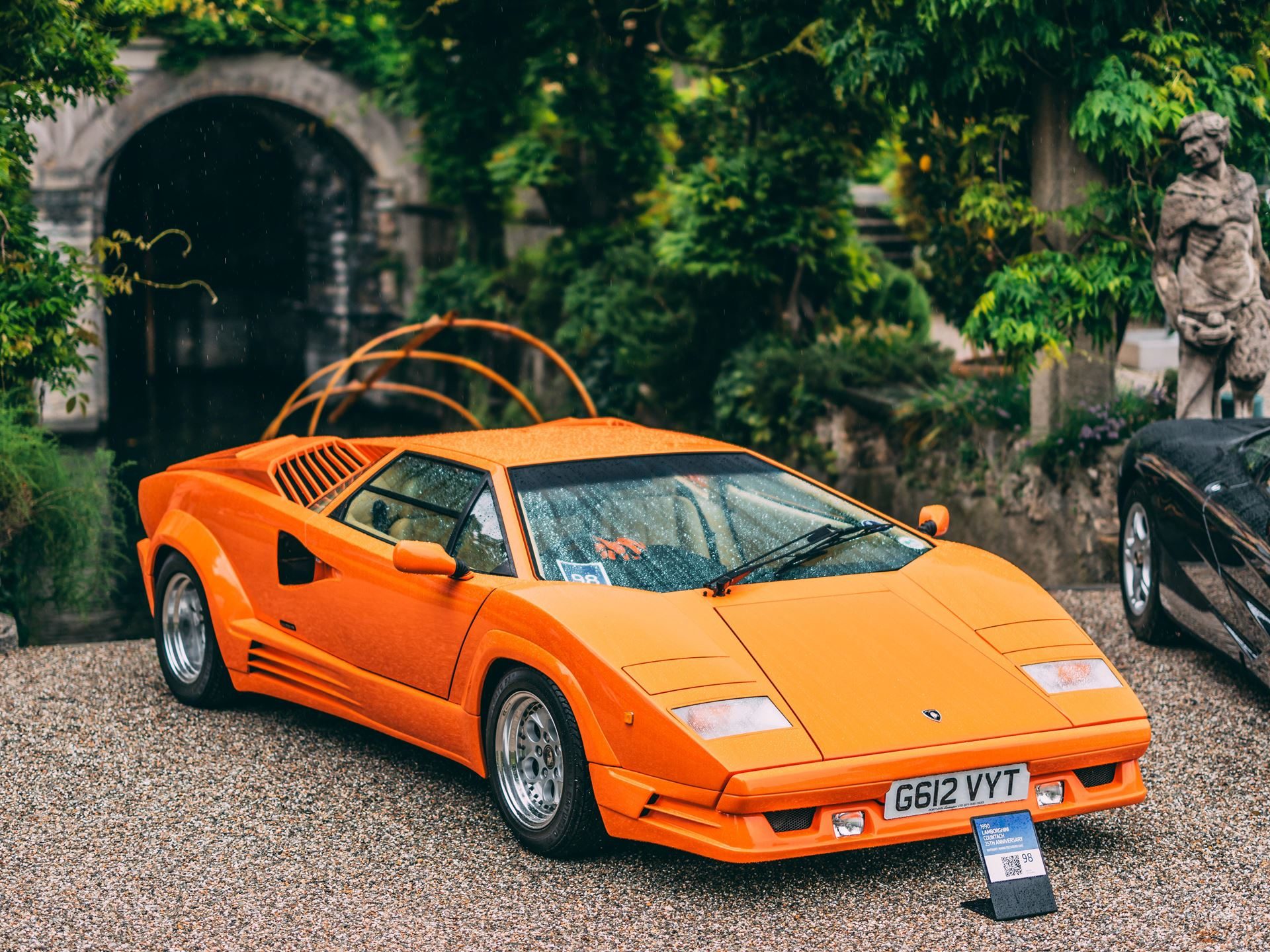 Orange lamborghini countach parked on gravel with classical architecture and greenery in the background