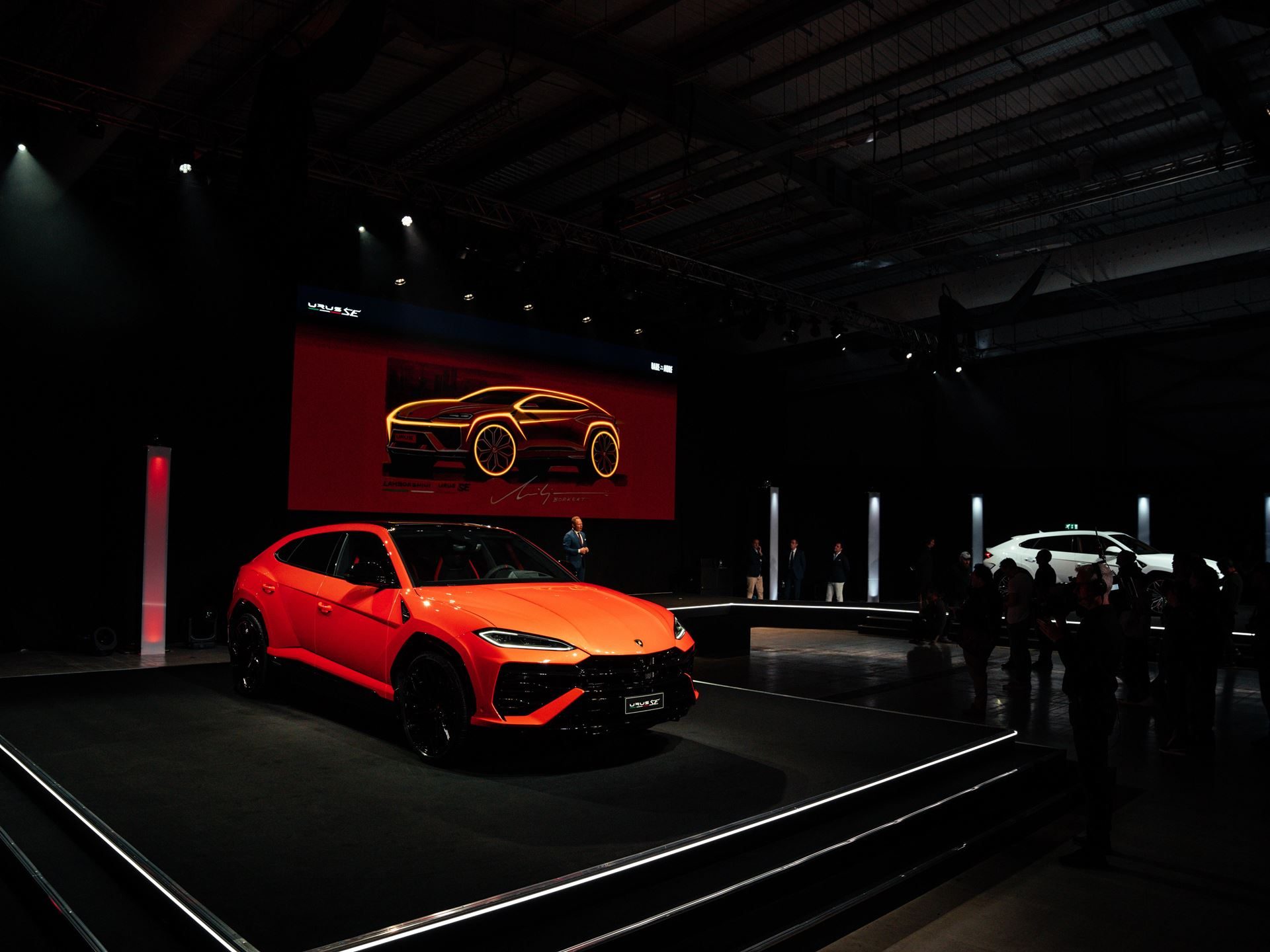 Lamborghini Day Uk Turned A Hybrid Reveal Into A Full Brand Experience, And That Was The Point 13 Orange lamborghini urus se on stage with a design sketch on a large screen and a white urus se visible in the background at lamborghini day uk 2024