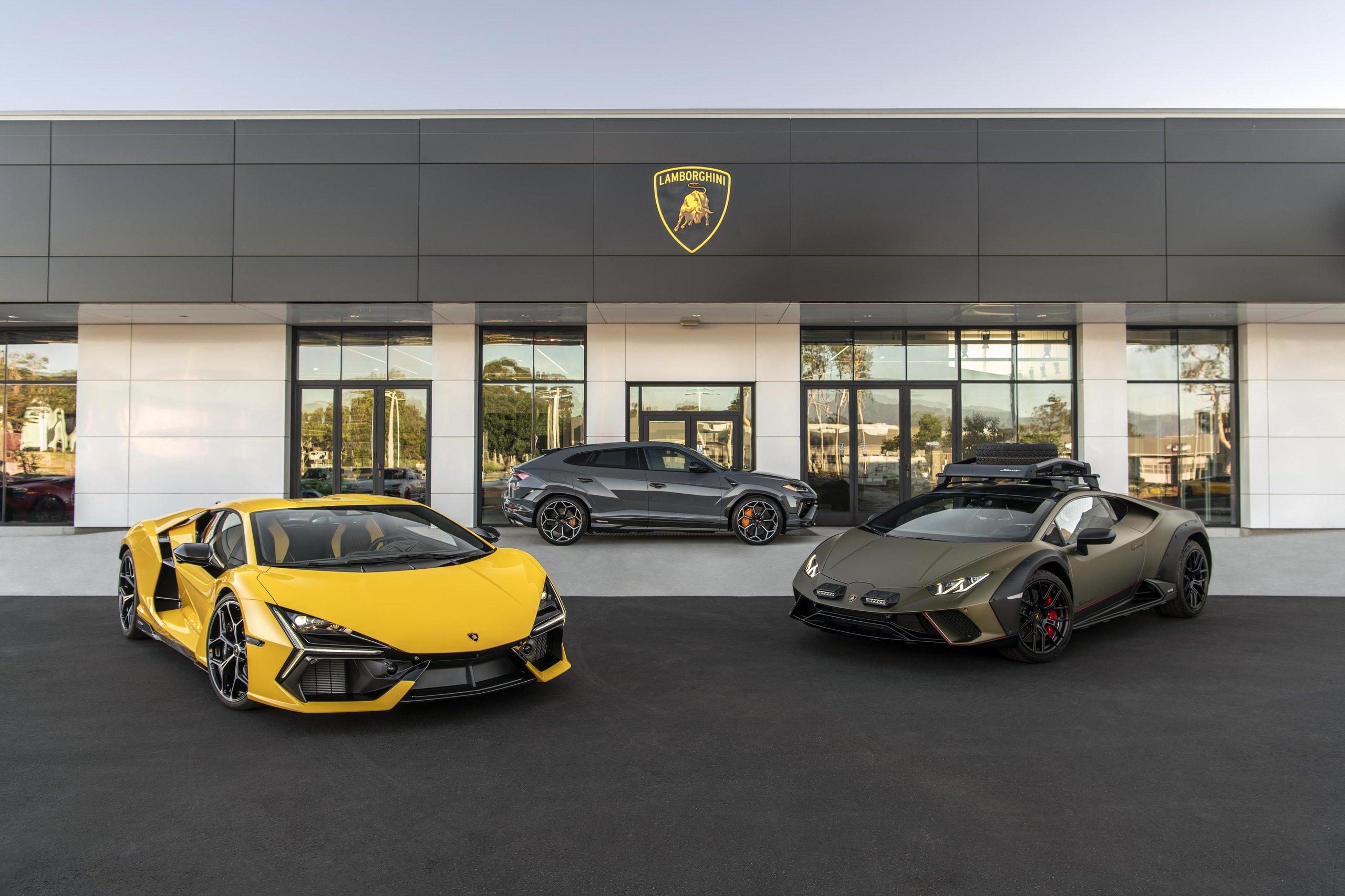 Six New U.s. Dealerships And Record Sales: How Lamborghini Built The Stage For Its Hybrid Era 3 A yellow revuelto, grey urus, and matte green huracán sterrato parked outside a modern lamborghini dealership