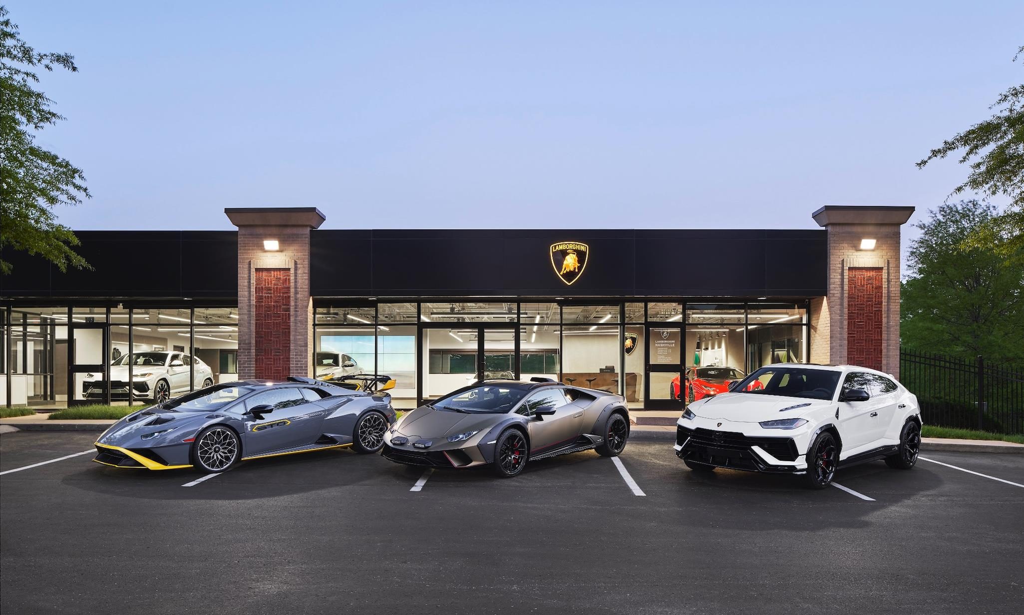 Six New U.s. Dealerships And Record Sales: How Lamborghini Built The Stage For Its Hybrid Era 5 A grey huracán tecnica, matte grey huracán sterrato, and white urus parked at dusk outside a lamborghini dealership