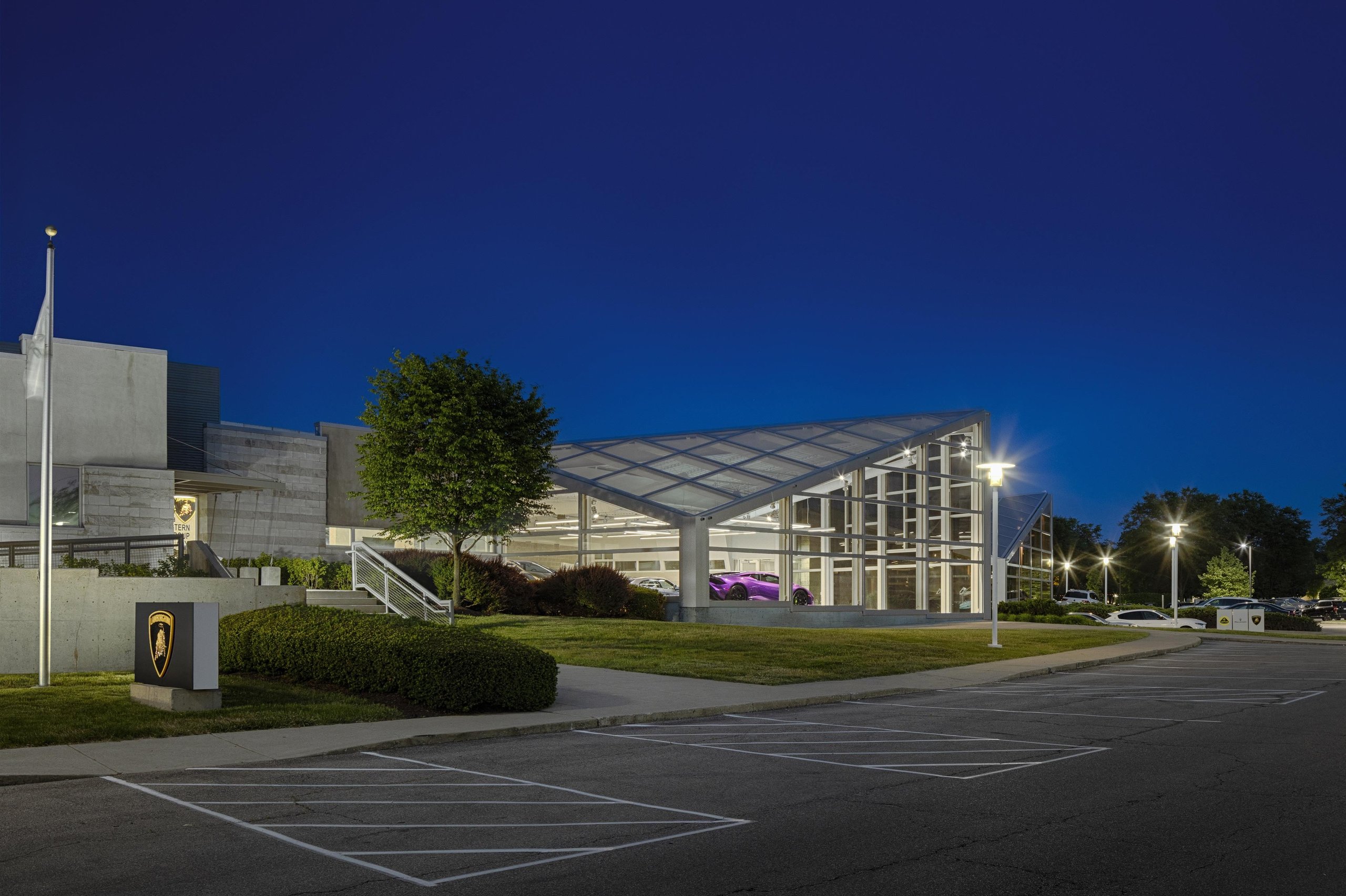 Six New U.s. Dealerships And Record Sales: How Lamborghini Built The Stage For Its Hybrid Era 9 Wide exterior view of a modern lamborghini dealership at dusk with a distinctive glass-roofed section and a purple huracán visible inside