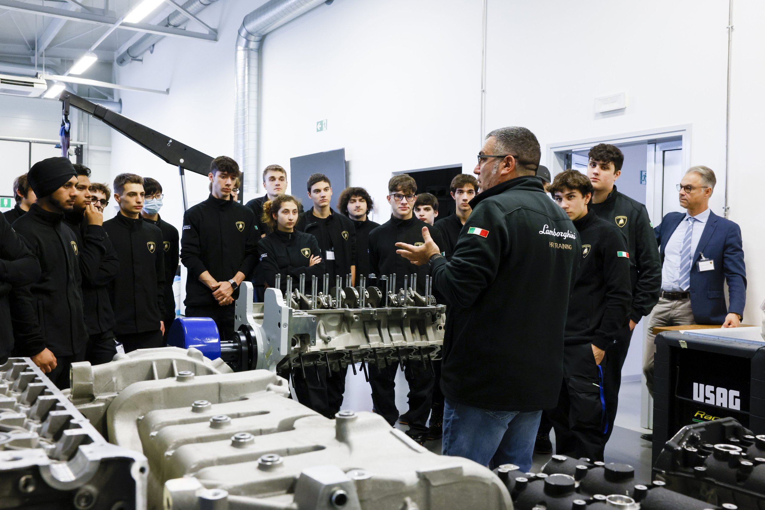 Inside Lamborghini'S Desi Program, Where 24 Students Train To Build Supercars 5 Lamborghini instructor explaining engine components to desi students in a workshop with disassembled engine parts visible on tables