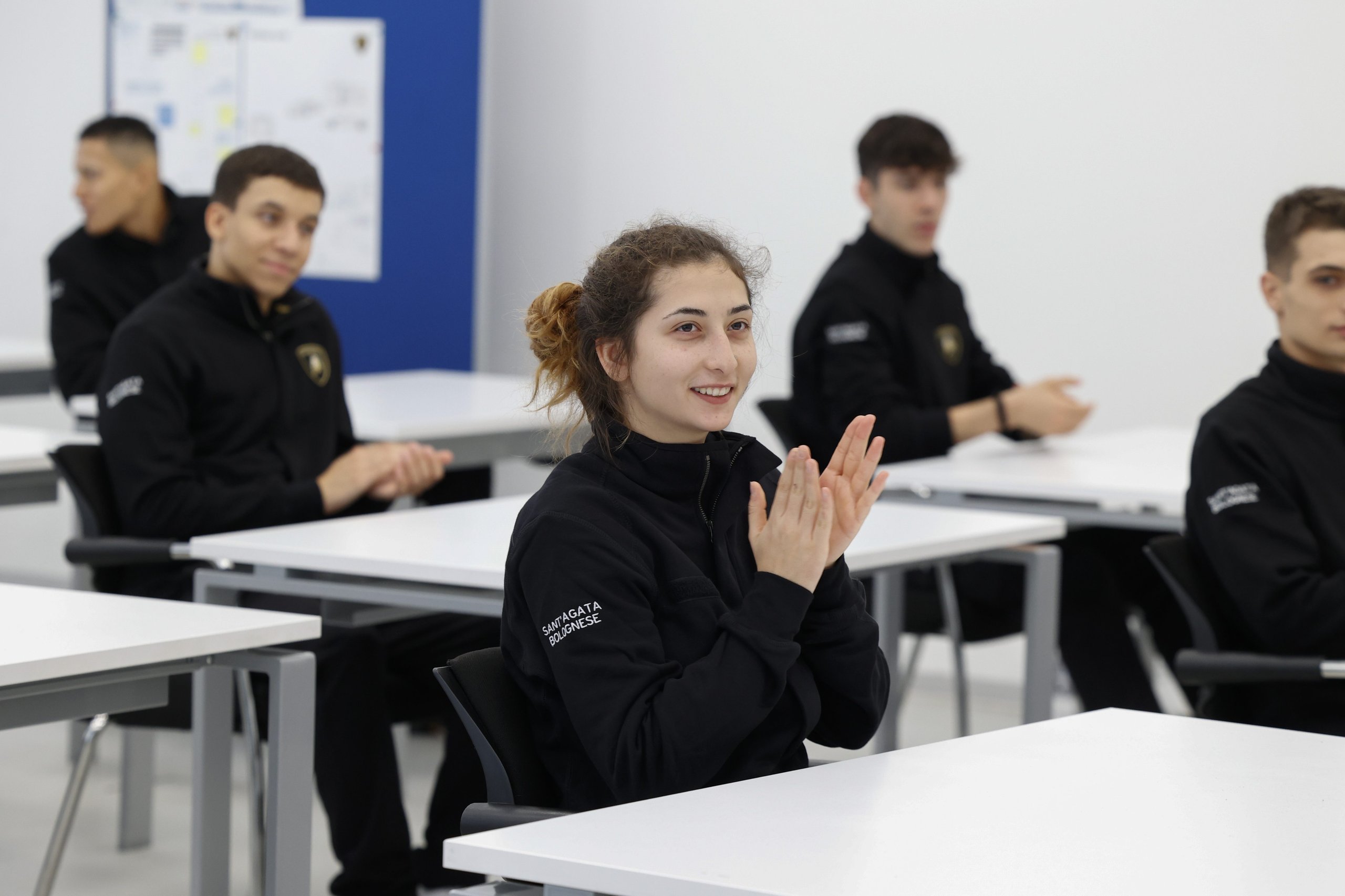 Inside Lamborghini'S Desi Program, Where 24 Students Train To Build Supercars 7 Female desi student in a lamborghini branded jacket clapping enthusiastically in a classroom alongside fellow students