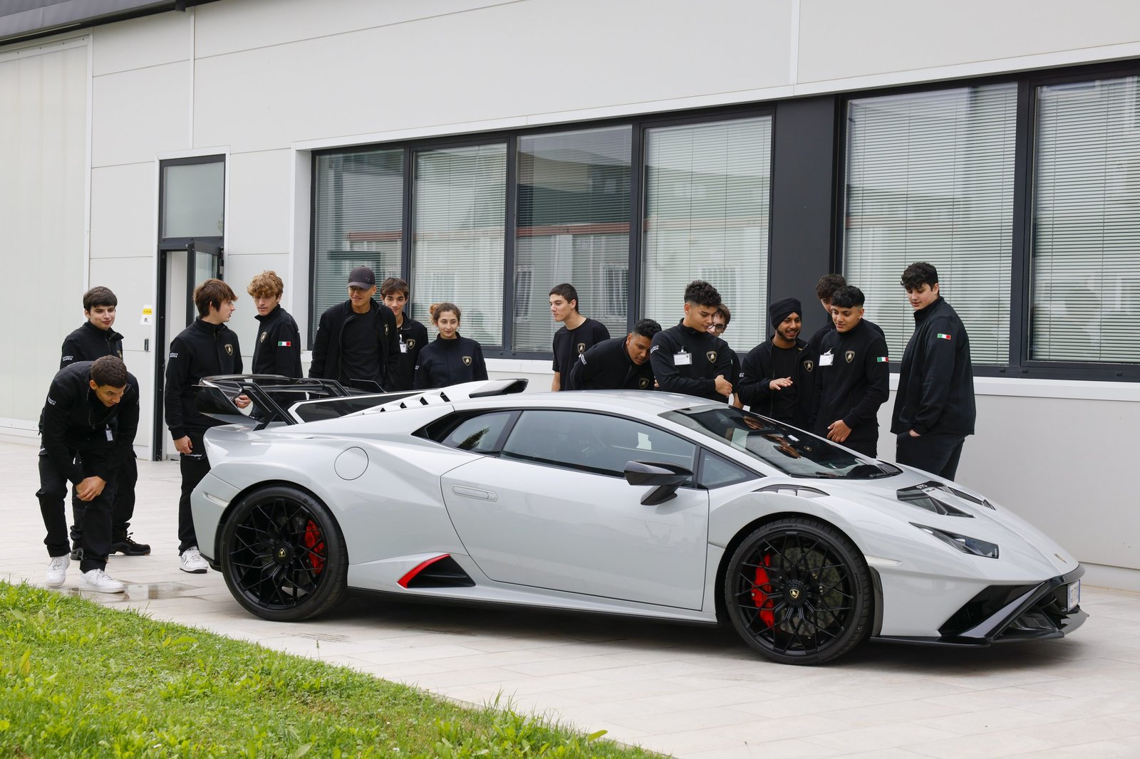 DESI students in Lamborghini HR Training jackets gathered around a grey Lamborghini Huracán outside the Sant'Agata Bolognese headquarters