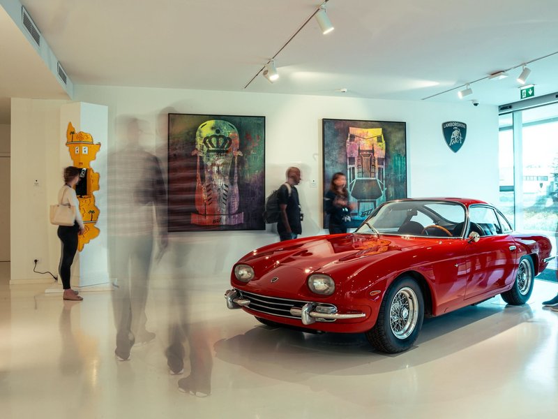 A classic red Lamborghini 350 GT displayed alongside contemporary artworks in the brightly lit Dreamaway exhibition hall at Sant'Agata Bolognese