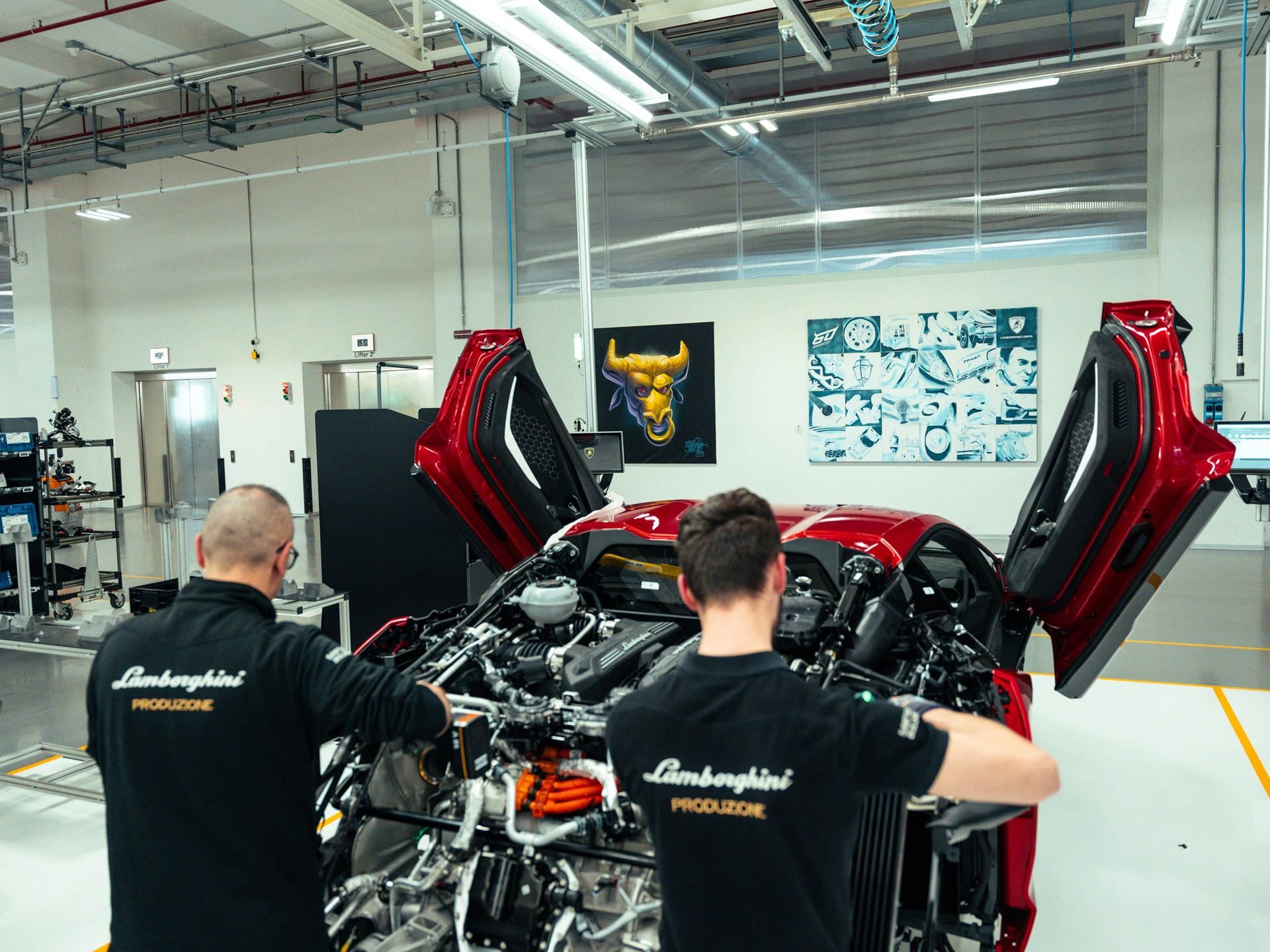 Two lamborghini production technicians work on a revuelto's exposed hybrid powertrain in the factory, with commissioned artworks visible on the wall behind them