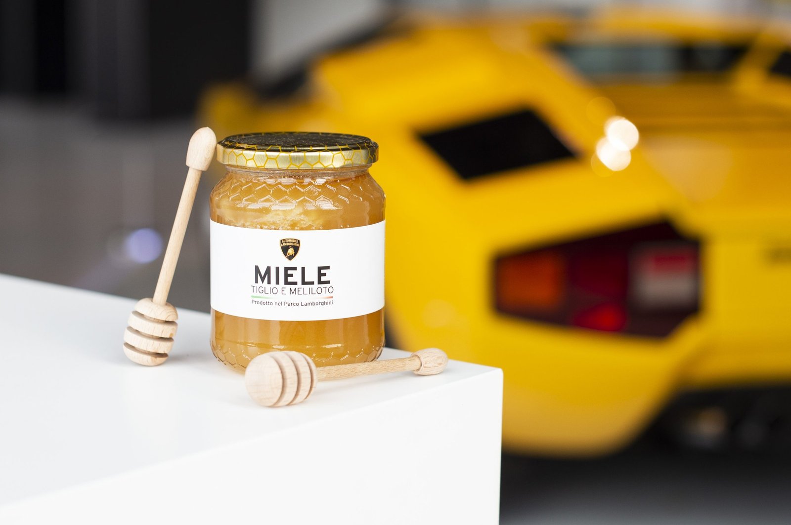 A jar of Lamborghini-branded honey with wooden dippers, a yellow Lamborghini visible in the background