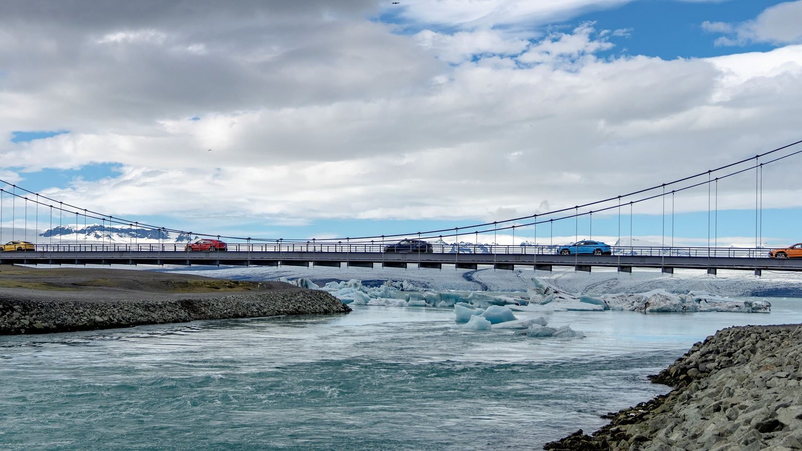 A convoy of colorful Lamborghini Urus SUVs crossing a suspension bridge over a glacial lagoon with icebergs and snow-capped mountains