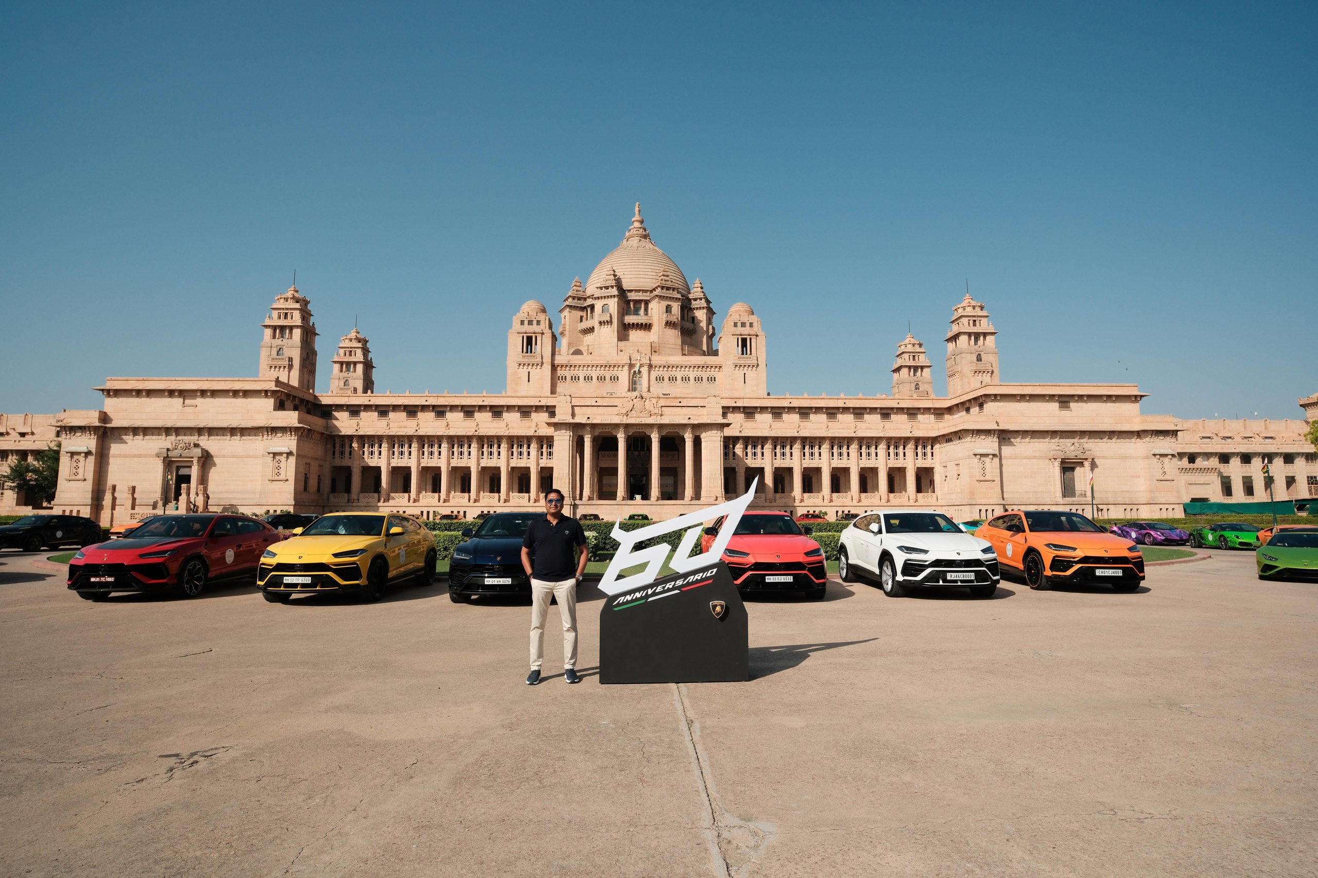 Lamborghini Esperienza Giro India 2023: A Royal 60Th Anniversary Celebration Across Rajasthan 9 Individual standing next to a 60th anniversary banner flanked by colorful lamborghini urus and huracán models with a grand palace in the background