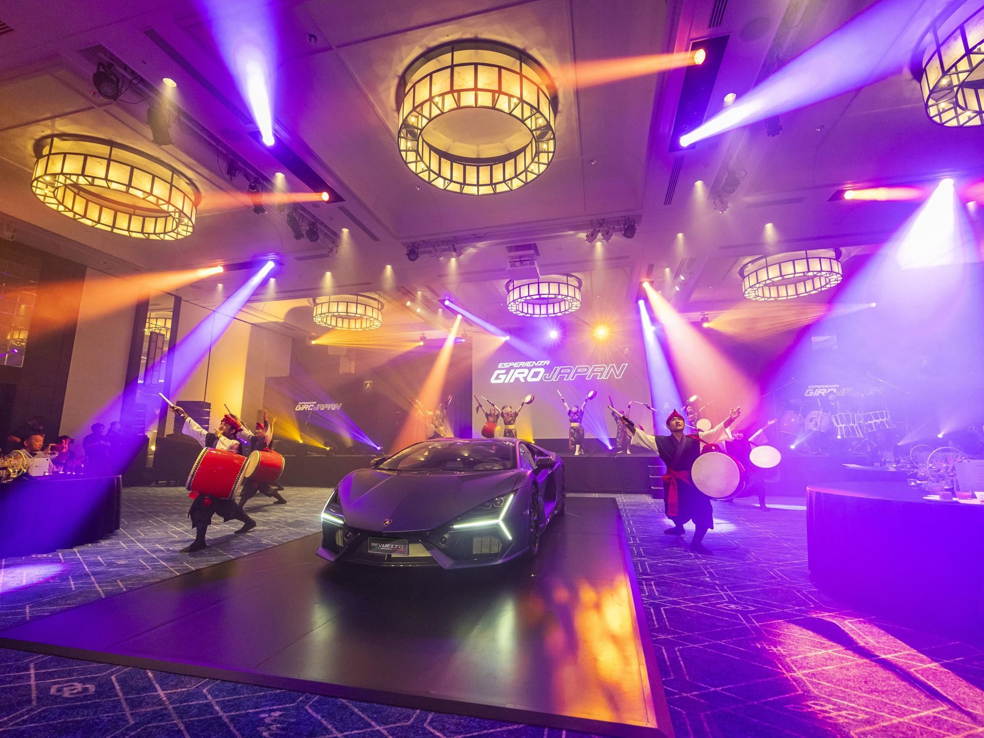 Lamborghini Turned Okinawa Into A Three-Day Supercar Convoy For Giro Japan 2024, With A Revuelto Debut As The Centerpiece 7 Dark grey lamborghini revuelto on an indoor stage surrounded by performers playing traditional japanese drums under vibrant stage lighting