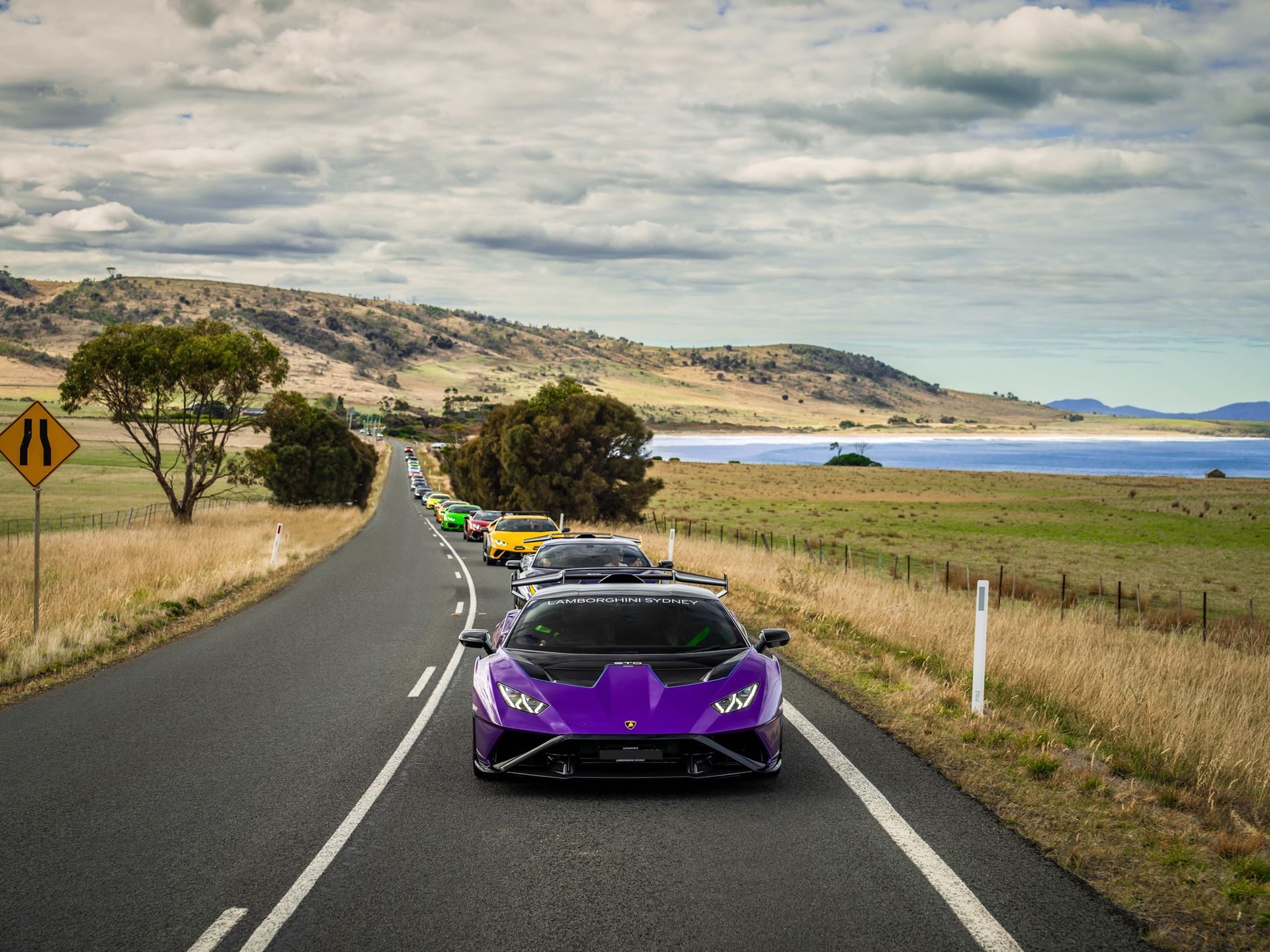 Lamborghini Took 40 Owners And 20 Supercars To Tasmania, And The Real Product Was The Trip Itself 3 A line of colorful lamborghinis led by a purple huracan sto driving down a winding rural tasmanian road with hills and water in the background