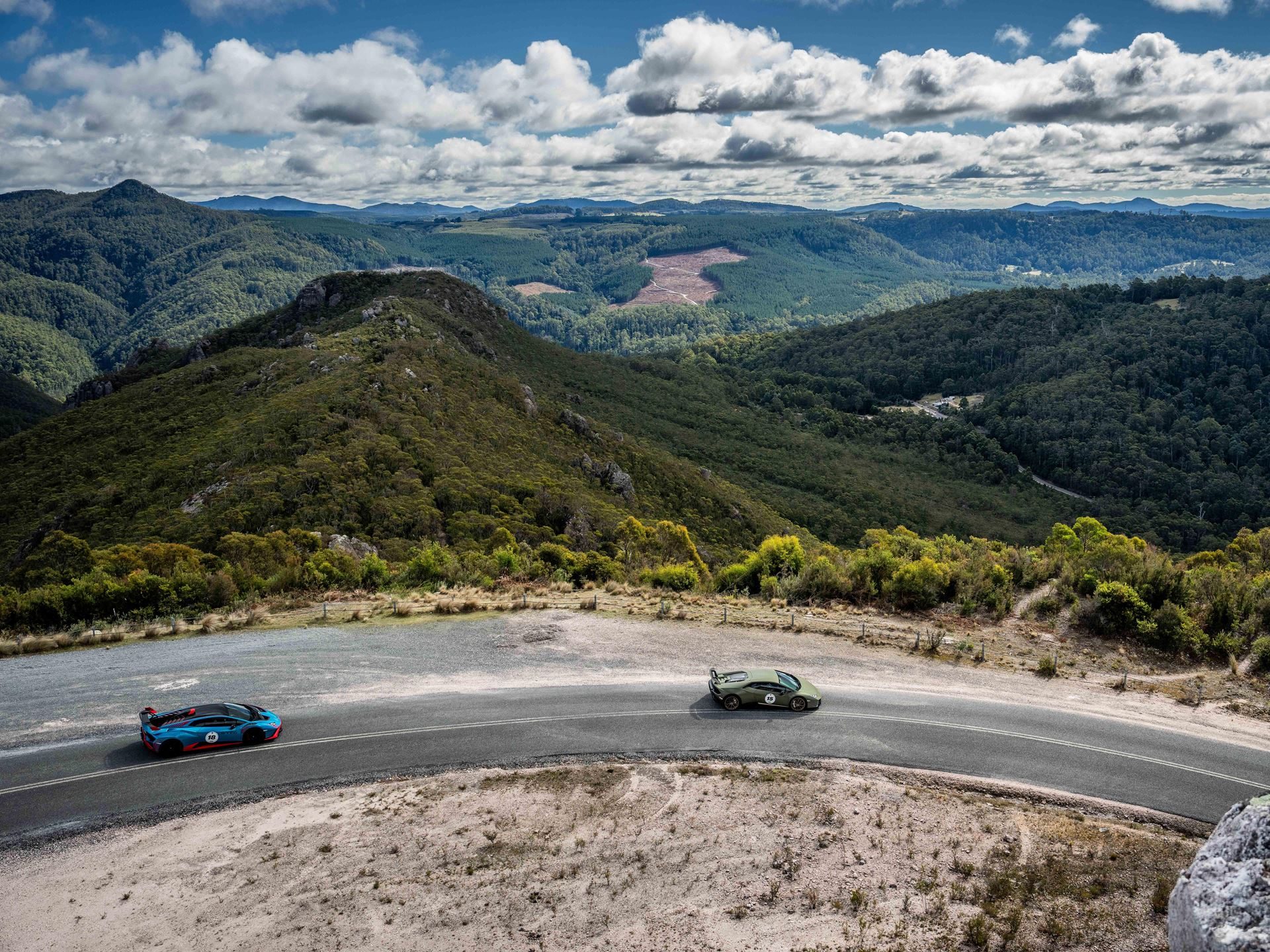 Lamborghini Took 40 Owners And 20 Supercars To Tasmania, And The Real Product Was The Trip Itself 7 A blue lamborghini huracán sto and a green urus driving together on a winding mountain road through lush green tasmanian hills