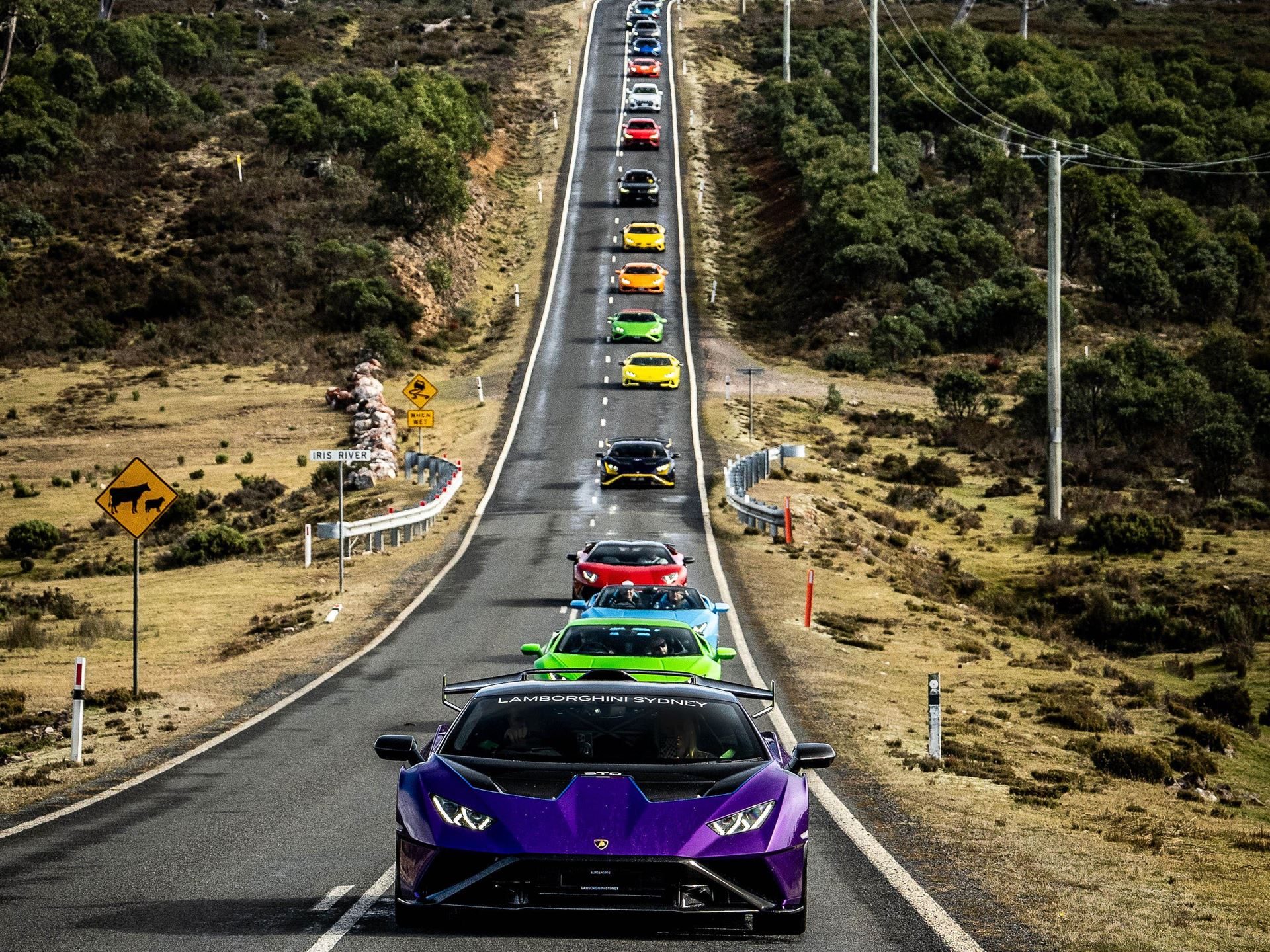 Lamborghini Took 40 Owners And 20 Supercars To Tasmania, And The Real Product Was The Trip Itself 9 A long line of various lamborghini models led by a purple huracán sto driving up a winding mountain road under a clear sky