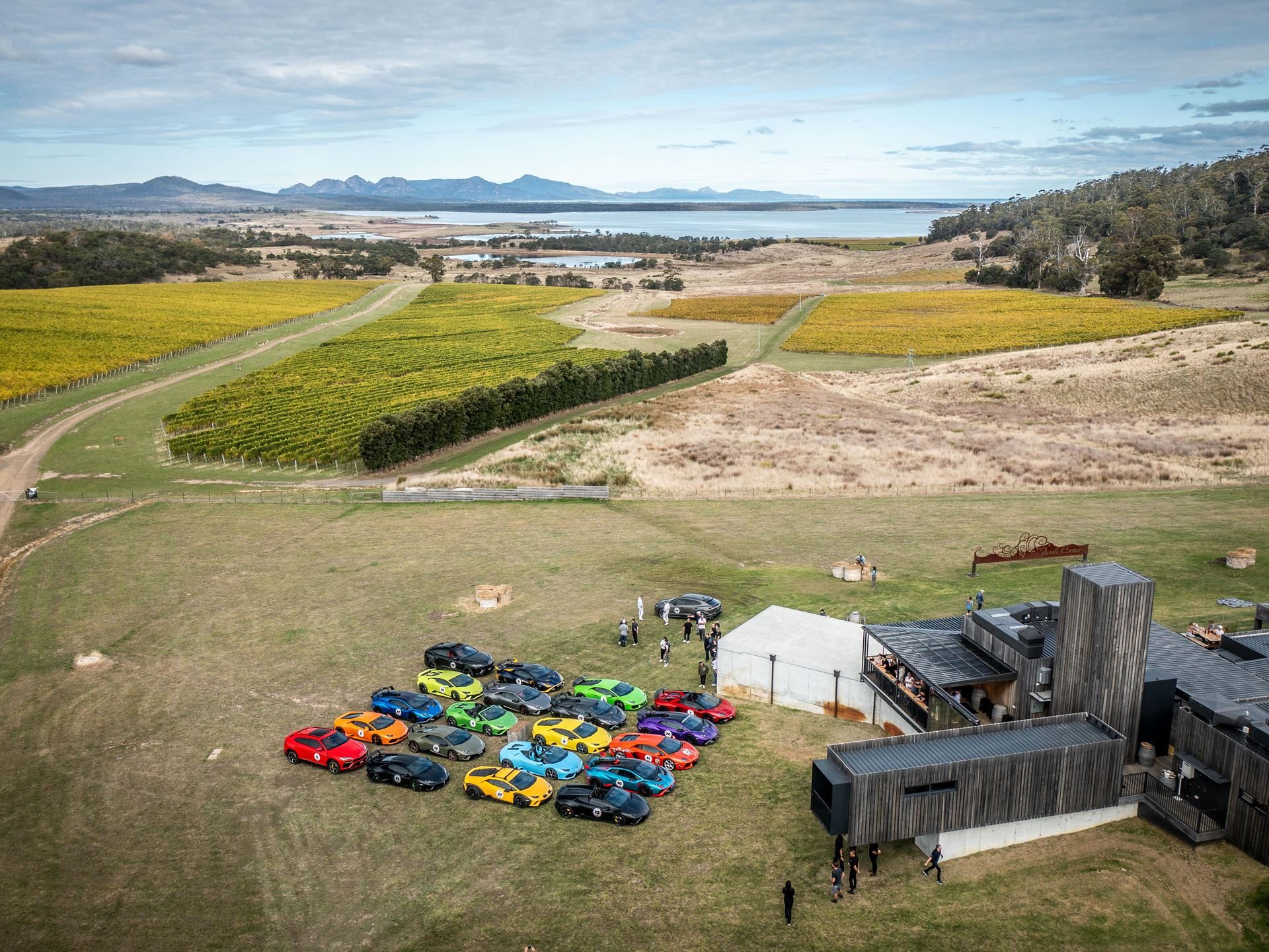 Lamborghini Took 40 Owners And 20 Supercars To Tasmania, And The Real Product Was The Trip Itself 5 Aerial view of colorful lamborghinis parked near a modern building surrounded by vineyards and a distant tasmanian coastline