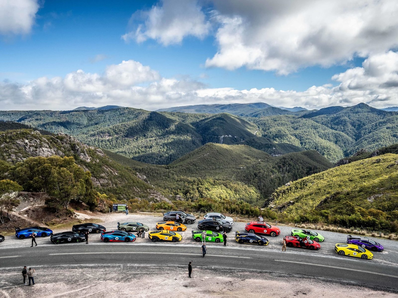 A large group of colorful Lamborghini supercars and SUVs parked at a mountain viewpoint overlooking Tasmania's green mountainous landscape