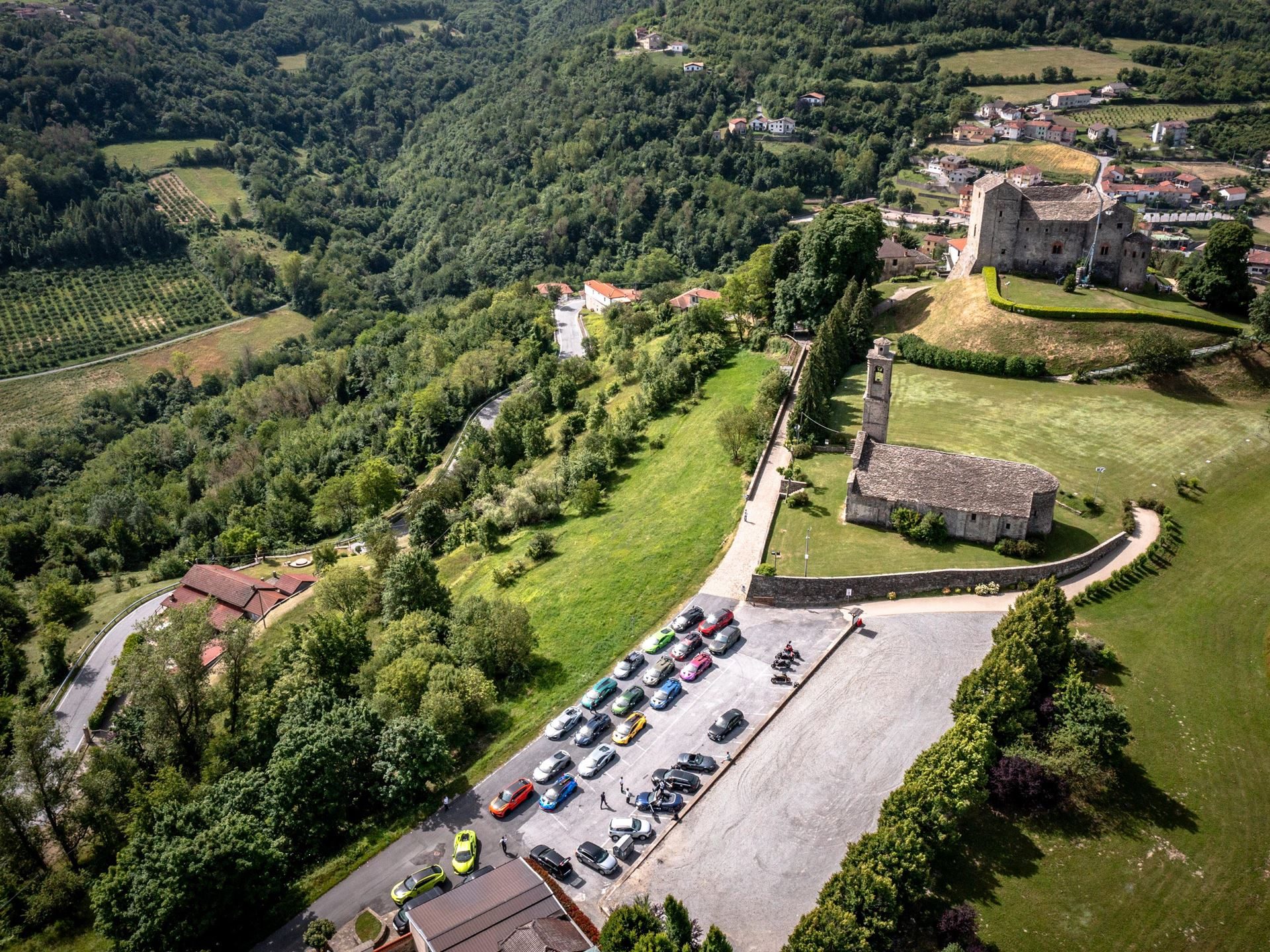 Aerial view of diverse lamborghini supercars parked next to a historic church and castle in a piedmont village