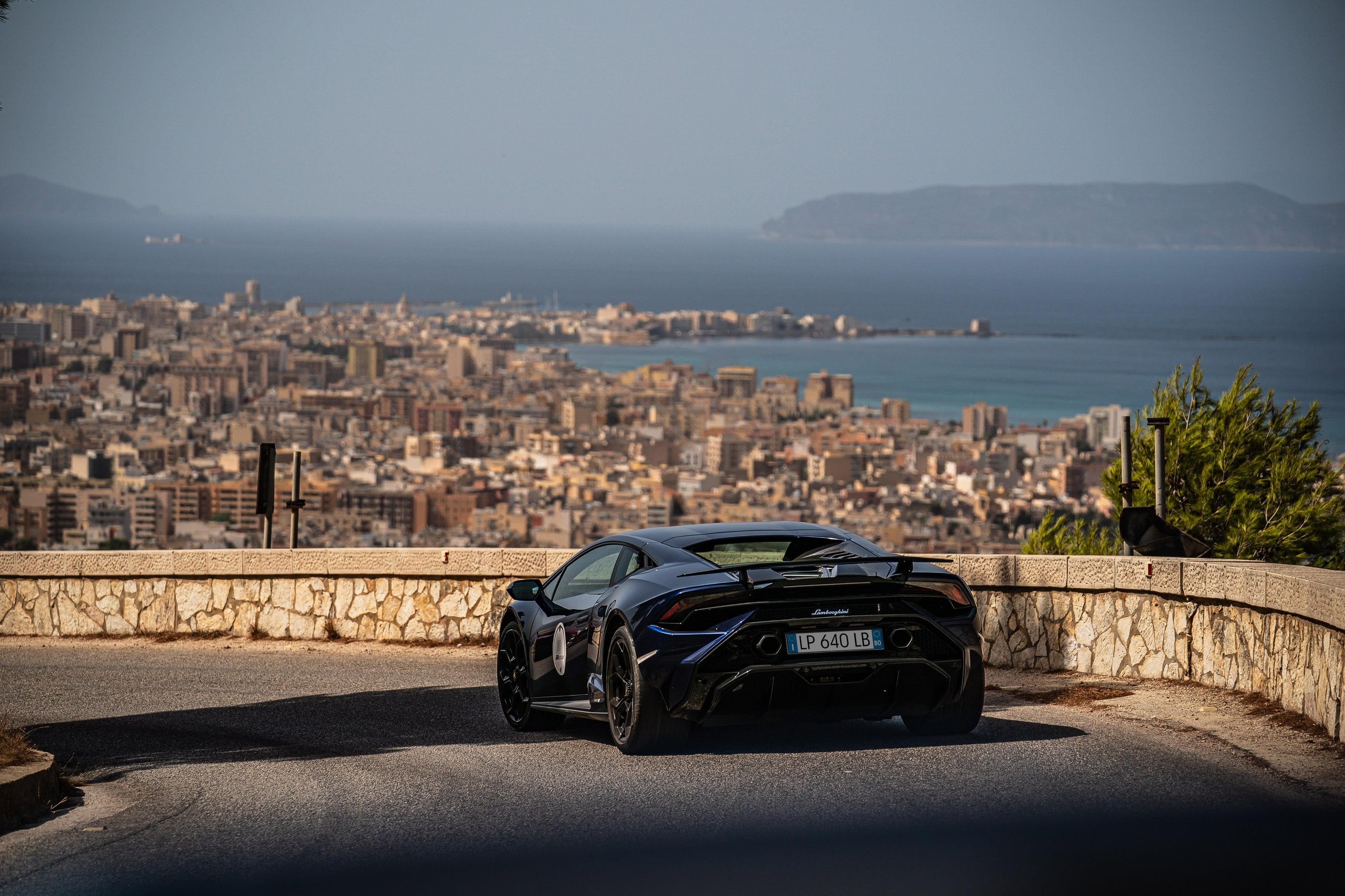 Esperienza Giro Sicilia: How Lamborghini Turned 750 Km Of Ancient Roads Into Its Most Personal Anniversary Statement 9 Dark blue lamborghini revuelto parked on a winding mountain road overlooking a sicilian coastal city and the mediterranean sea