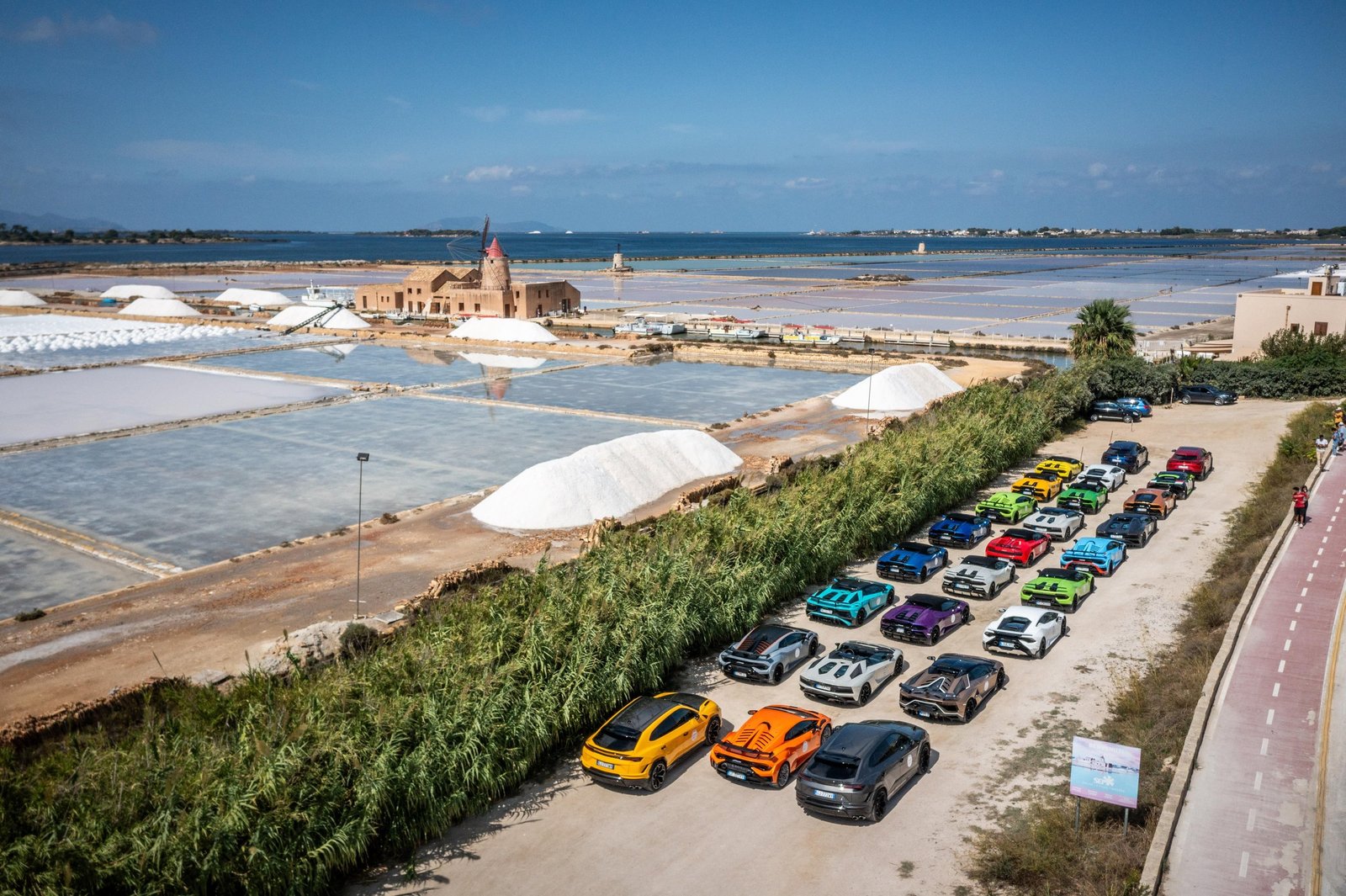 Aerial view of a colorful convoy of Lamborghini Huracán and Urus models parked alongside Sicilian salt flats with a historic building in the background