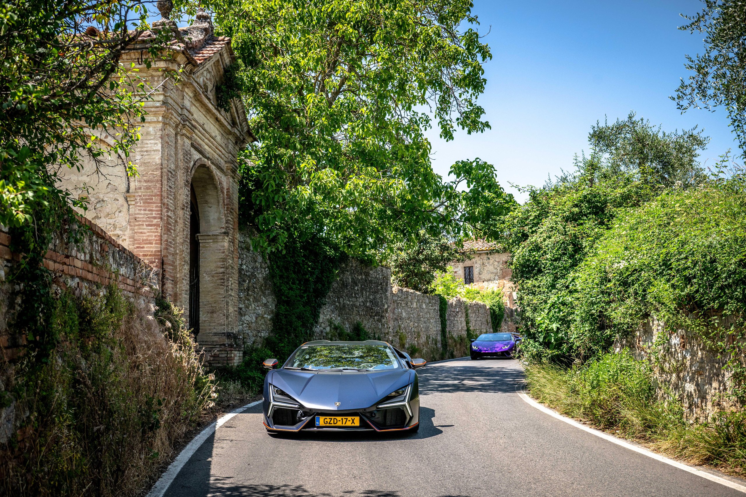 Two lamborghini sián fkp 37 supercars driving along a winding road through lush green tuscan landscape with ancient stone walls