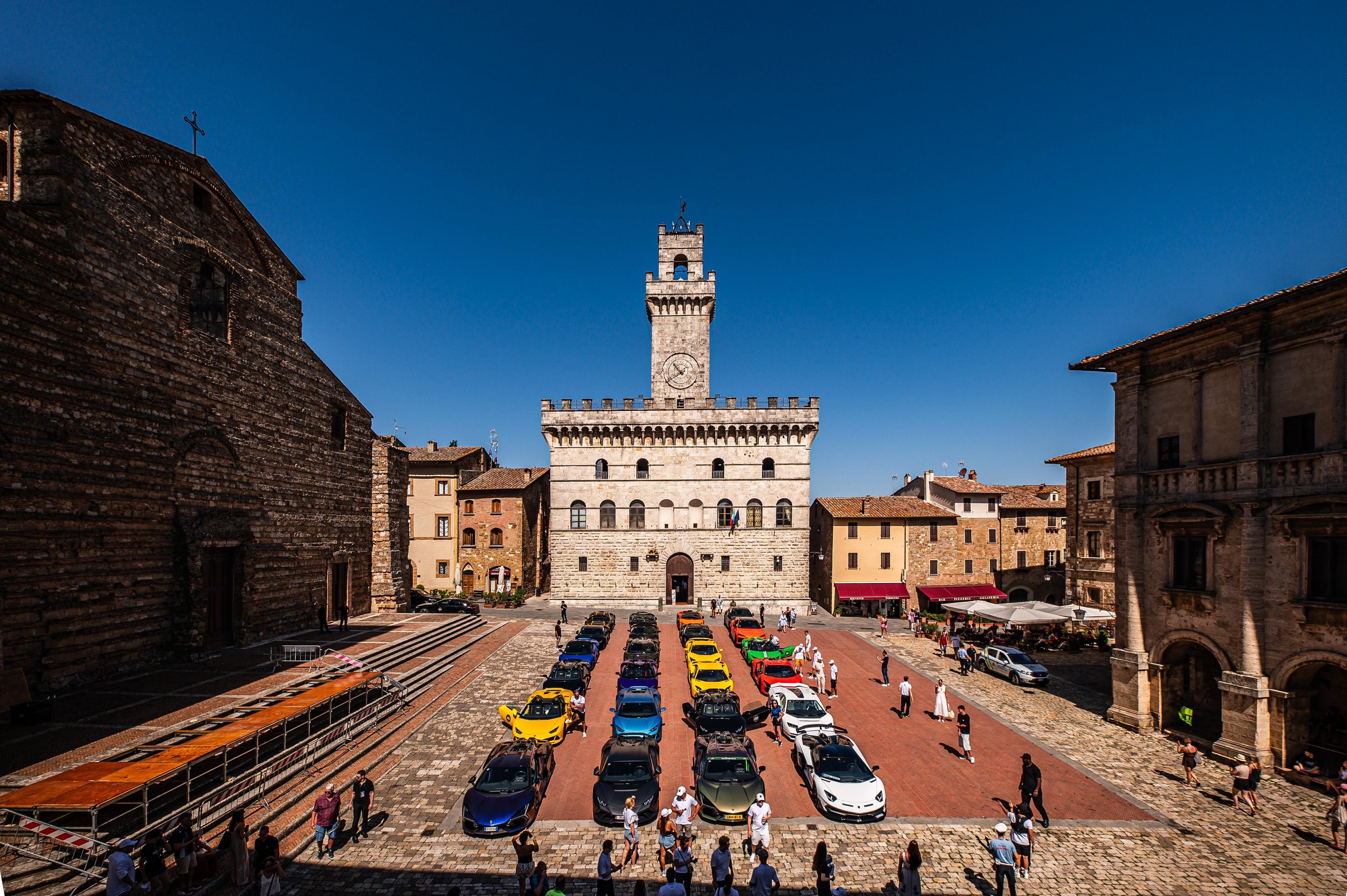 Historic italian town square filled with colorful lamborghini supercars and suvs with a clock tower in the background