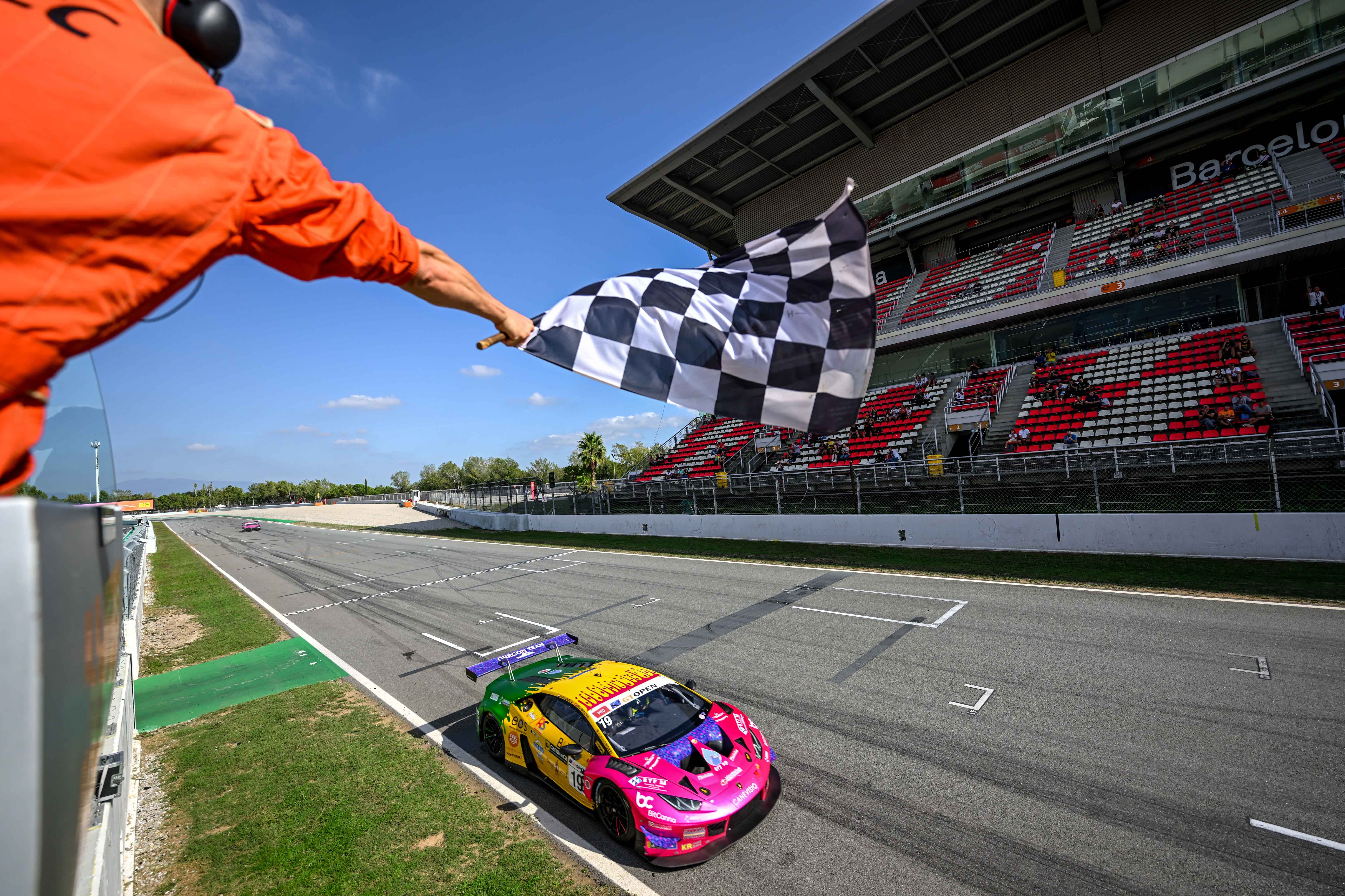 A Lamborghini Huracán GT3 Evo in vibrant green, yellow, and pink livery crosses the finish line under the checkered flag at Barcelona
