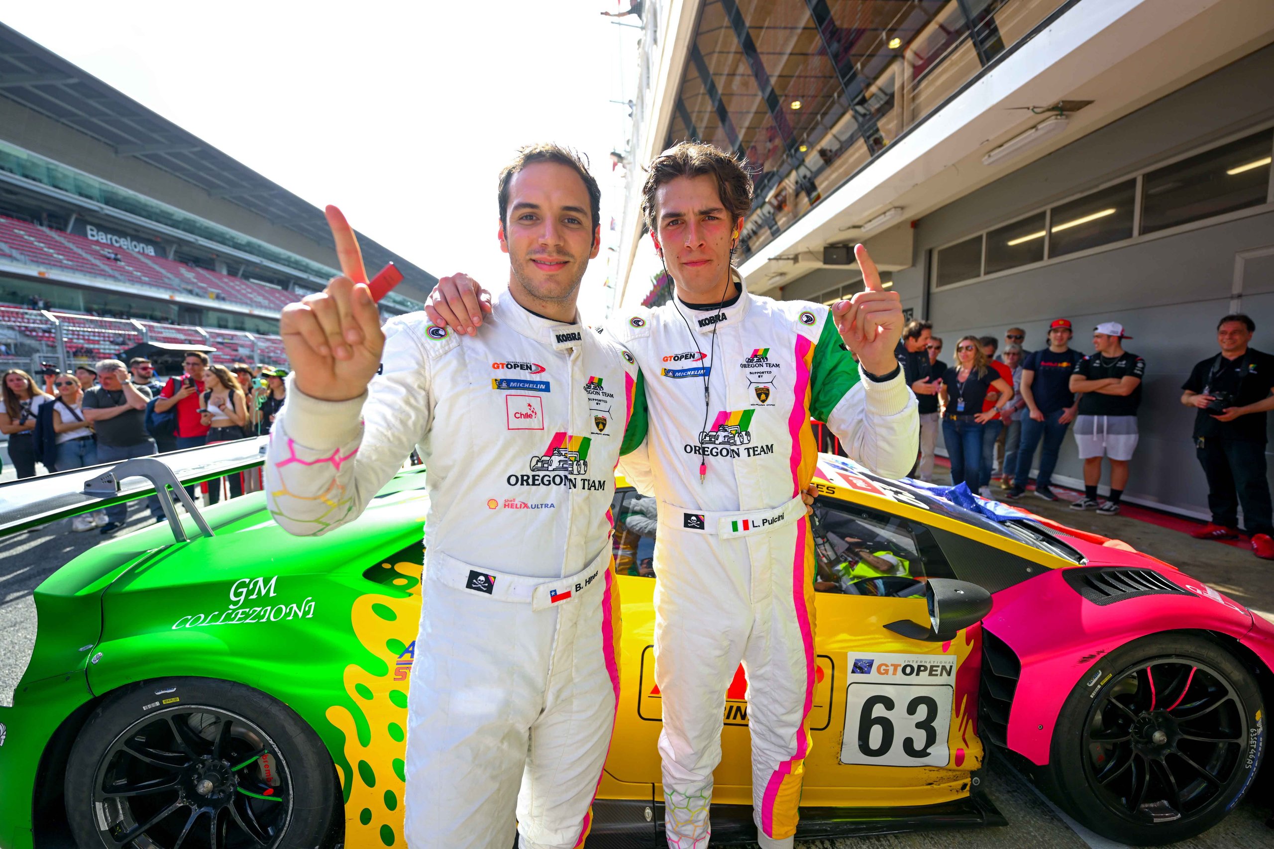 Two Oregon Team racing drivers in white suits stand alongside their brightly liveried Lamborghini Huracán GT3 Evo in the pit lane at Barcelona