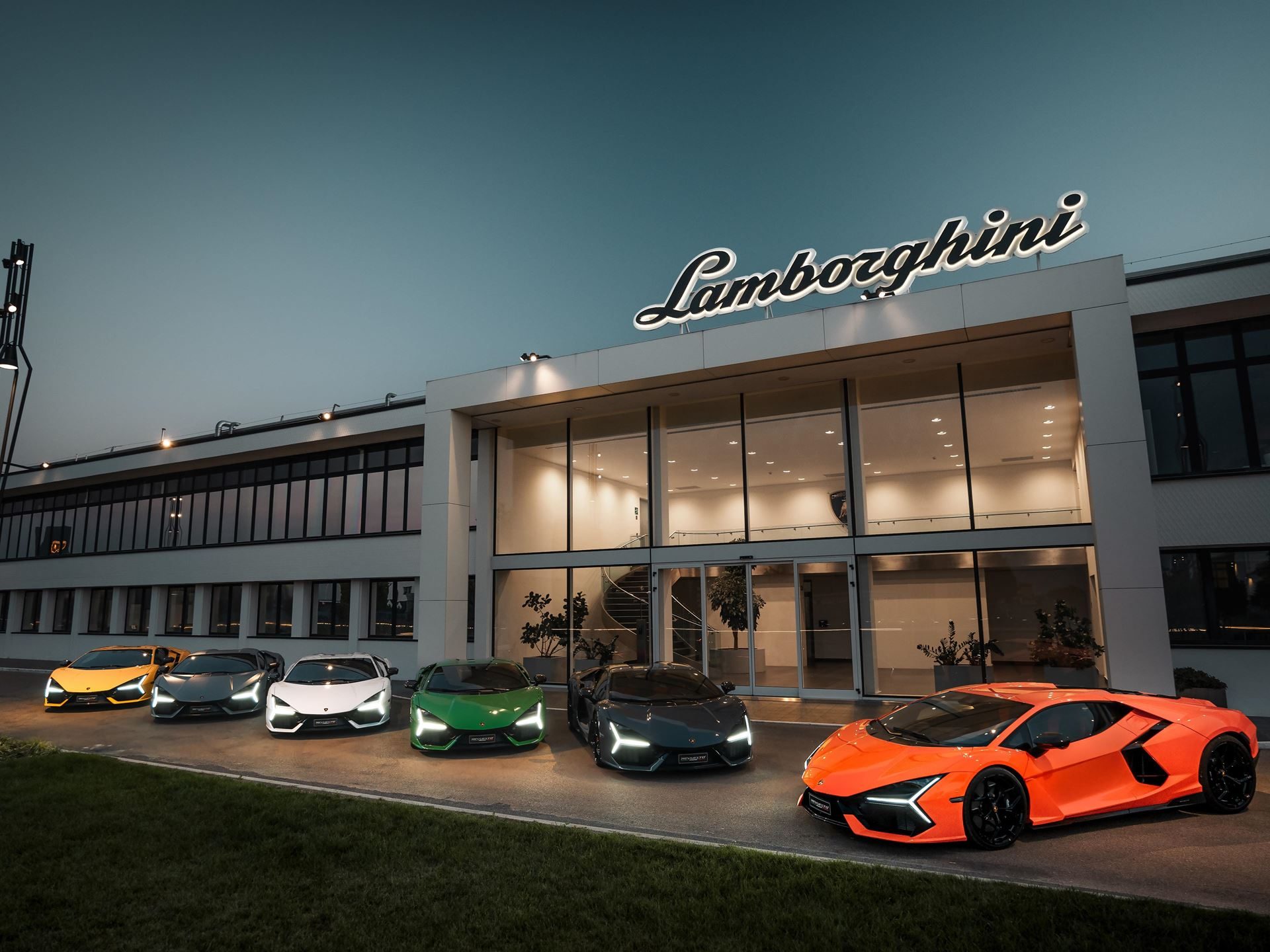Lineup of six lamborghini supercars including an orange revuelto in front of illuminated factory building at dusk