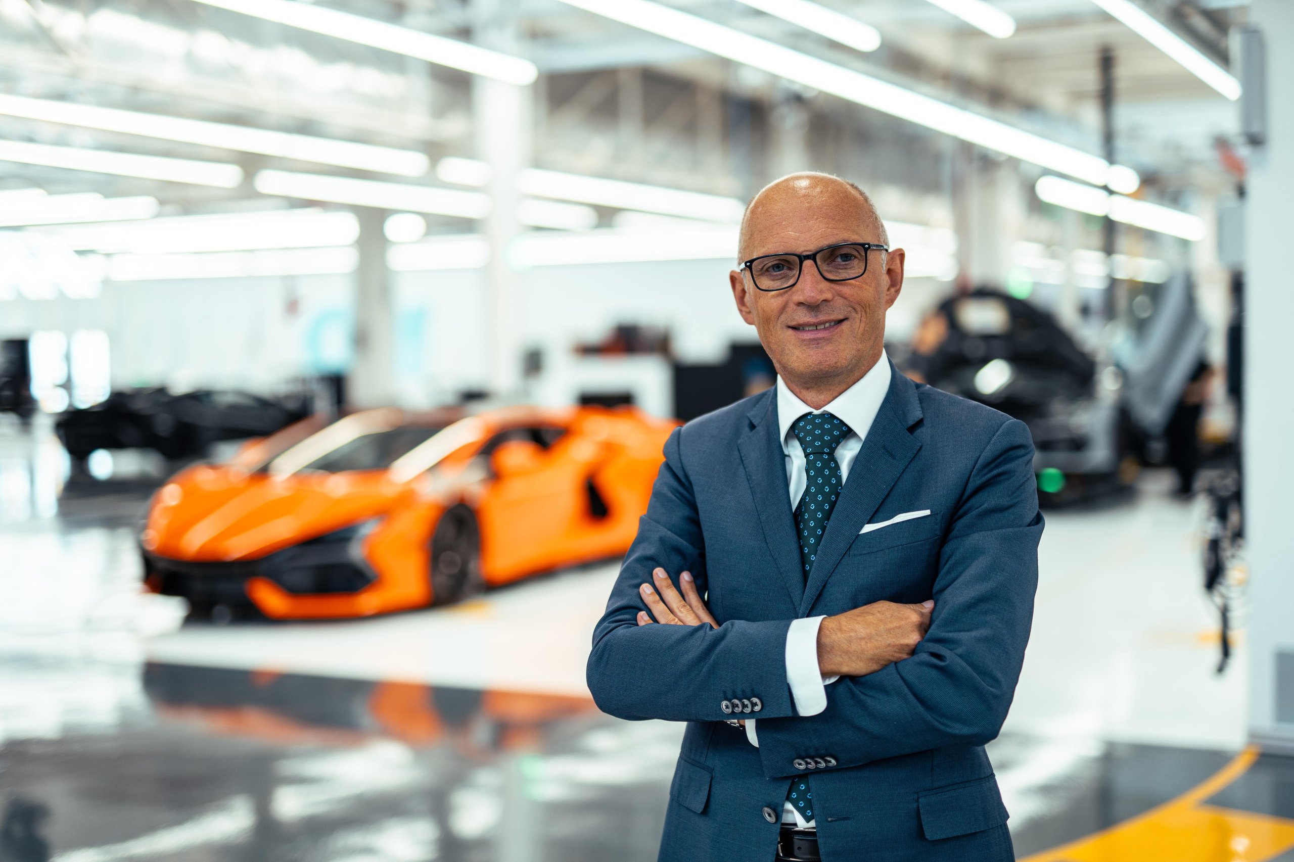 Lamborghini executive in suit standing on factory floor with orange revuelto in background