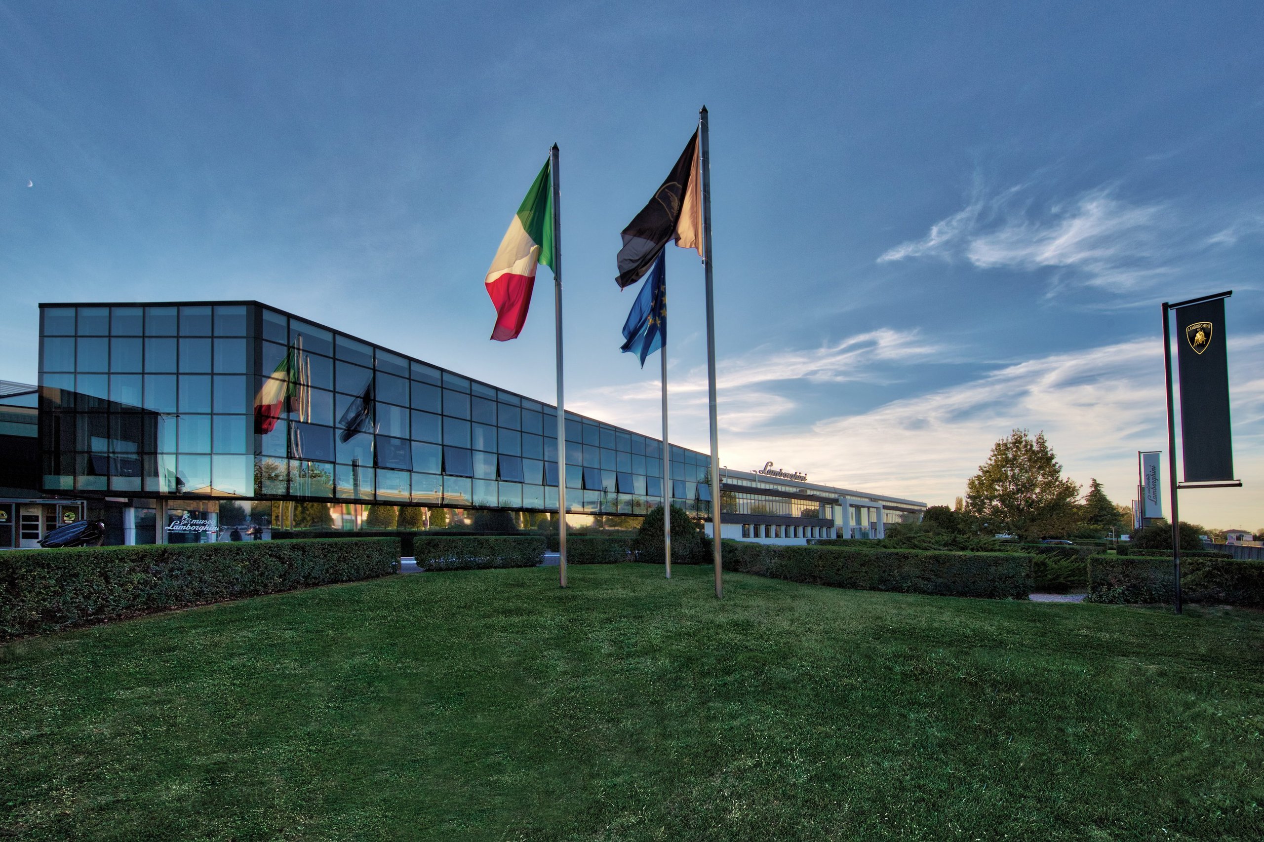 Lamborghini Puts One Executive In Charge Of Both Marketing And Sales, Hires Bugatti'S Comms Chief: Why It Matters 11 Lamborghini headquarters at dusk with flags flying against a colorful evening sky