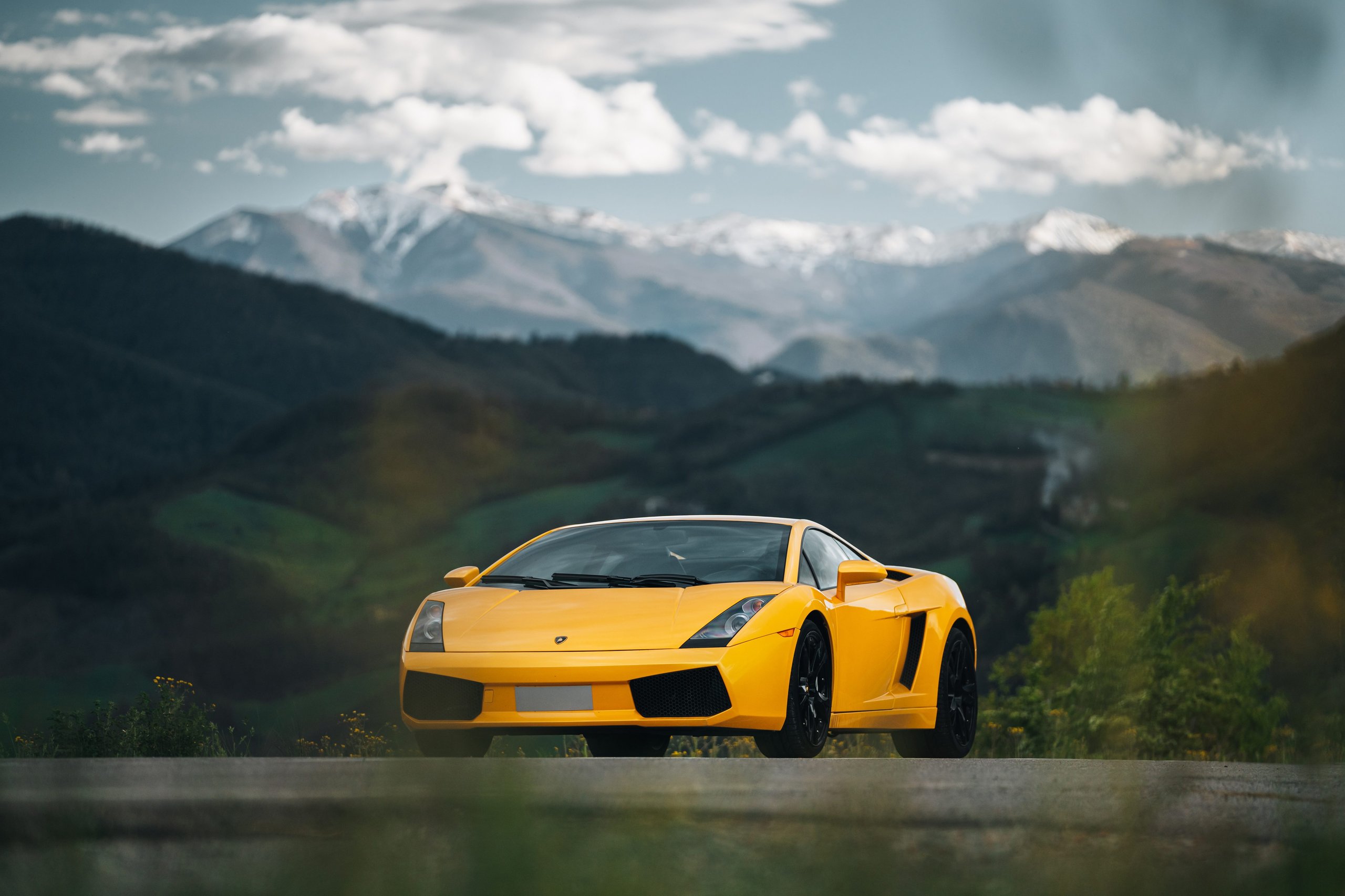 Yellow Lamborghini Gallardo parked on a road with snow-capped mountains and green hills in the background