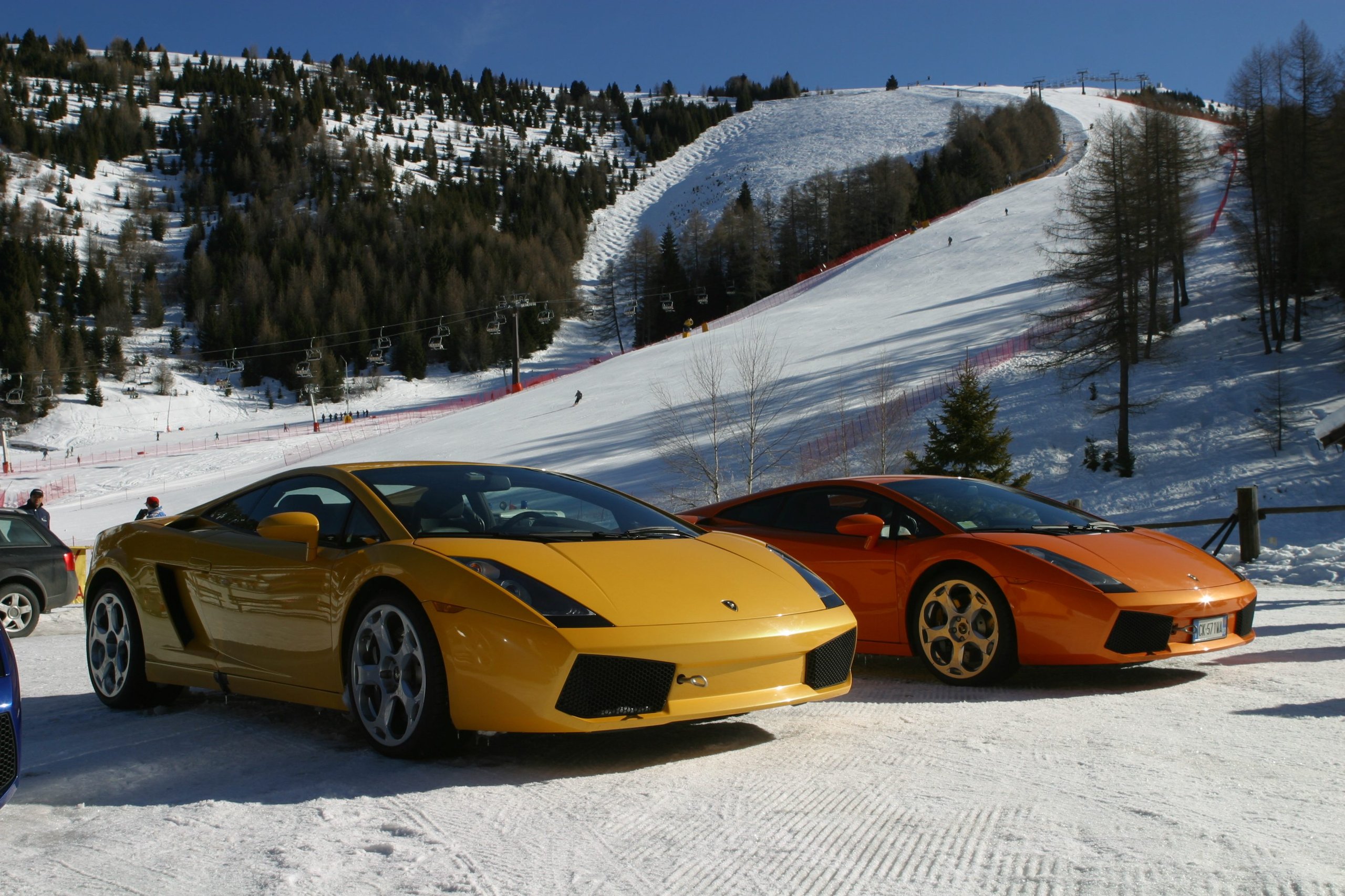Yellow and orange Lamborghini Gallardos parked on snow at a ski resort with slopes in the background