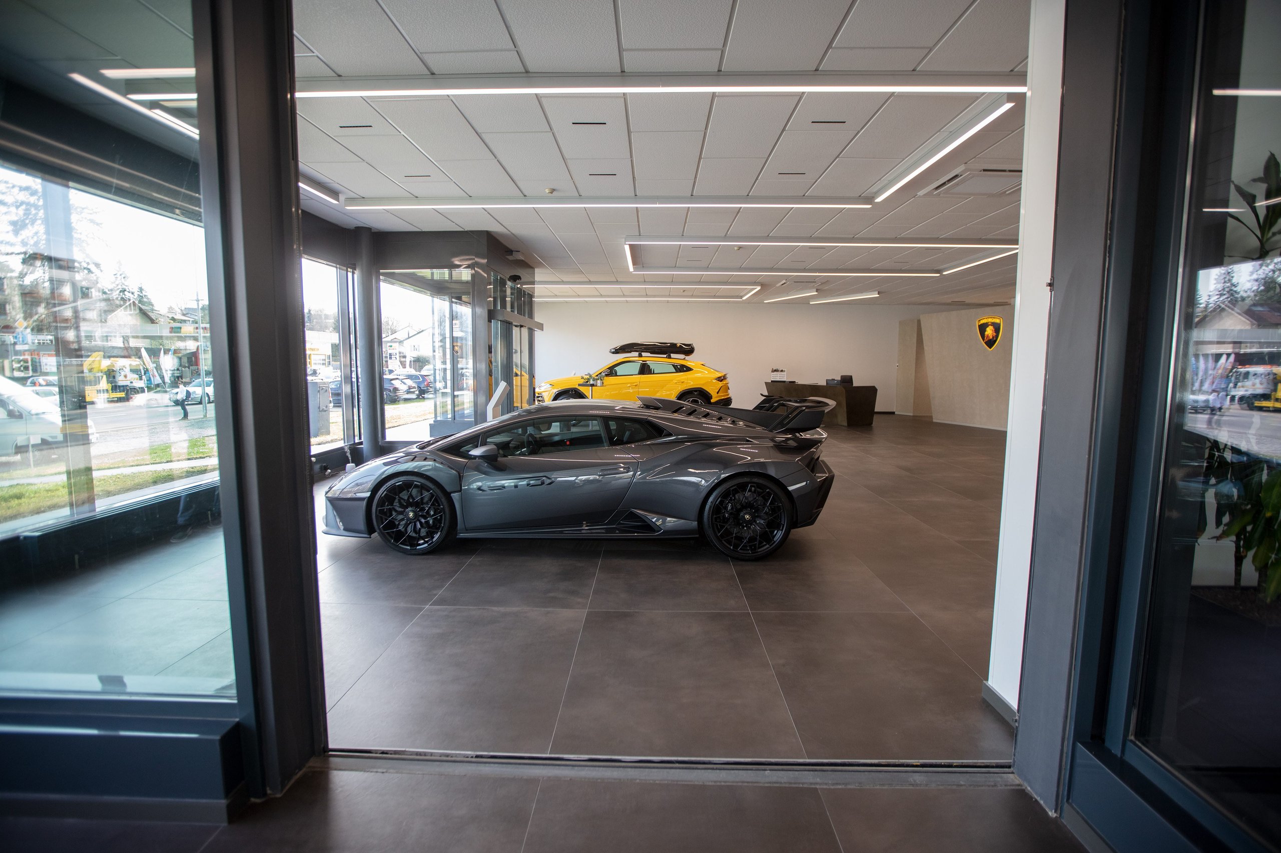 Lamborghini Geneva Celebrates 20 Years With A Showroom Built For The Brand'S Next Chapter 3 Grey lamborghini huracán sto and yellow lamborghini urus displayed inside the modern geneva showroom viewed through a large glass entrance