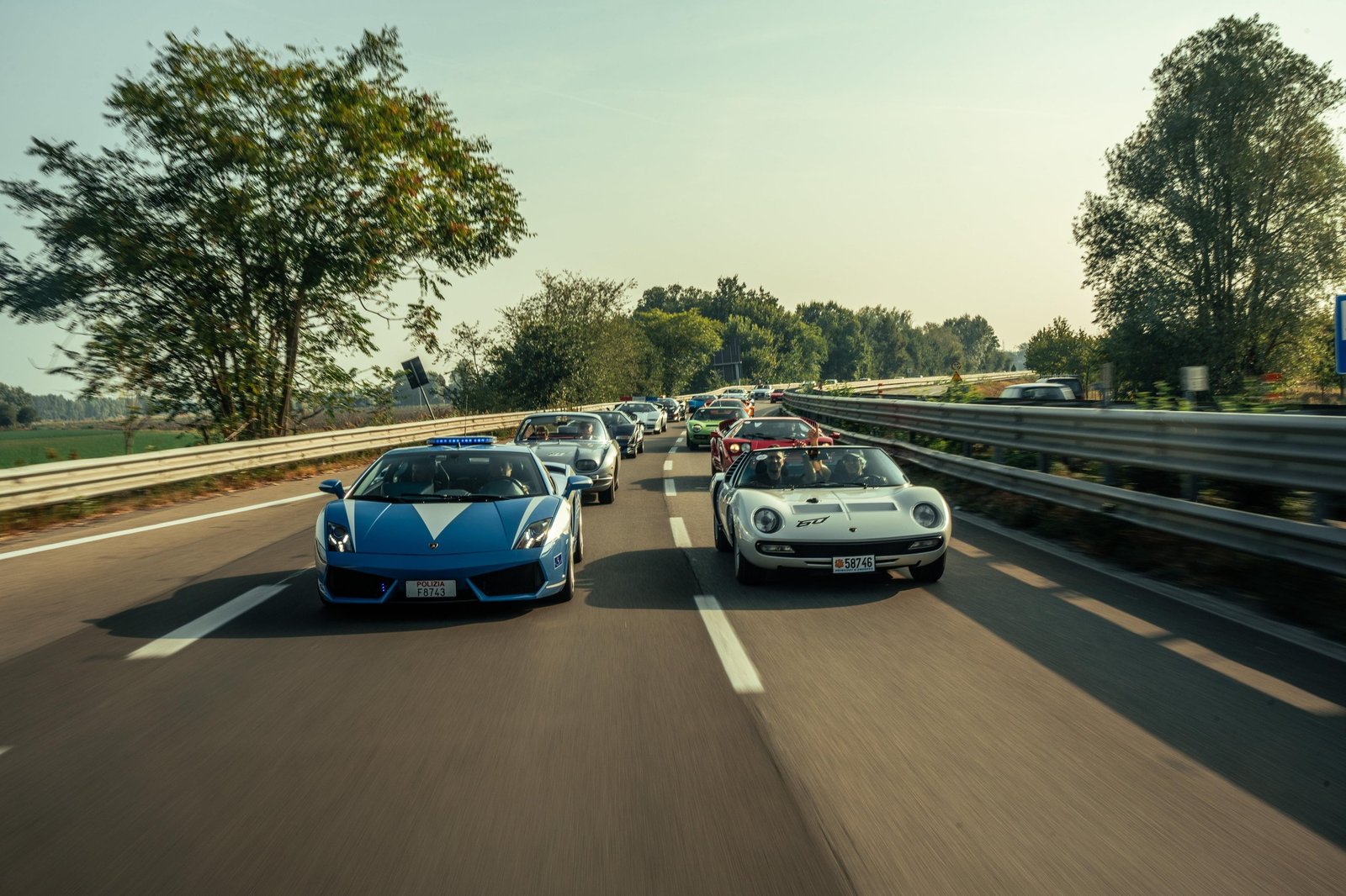 A convoy of classic Lamborghinis including a white Miura follows a blue and white Gallardo police car on an Italian highway during the Giro Polo Storico