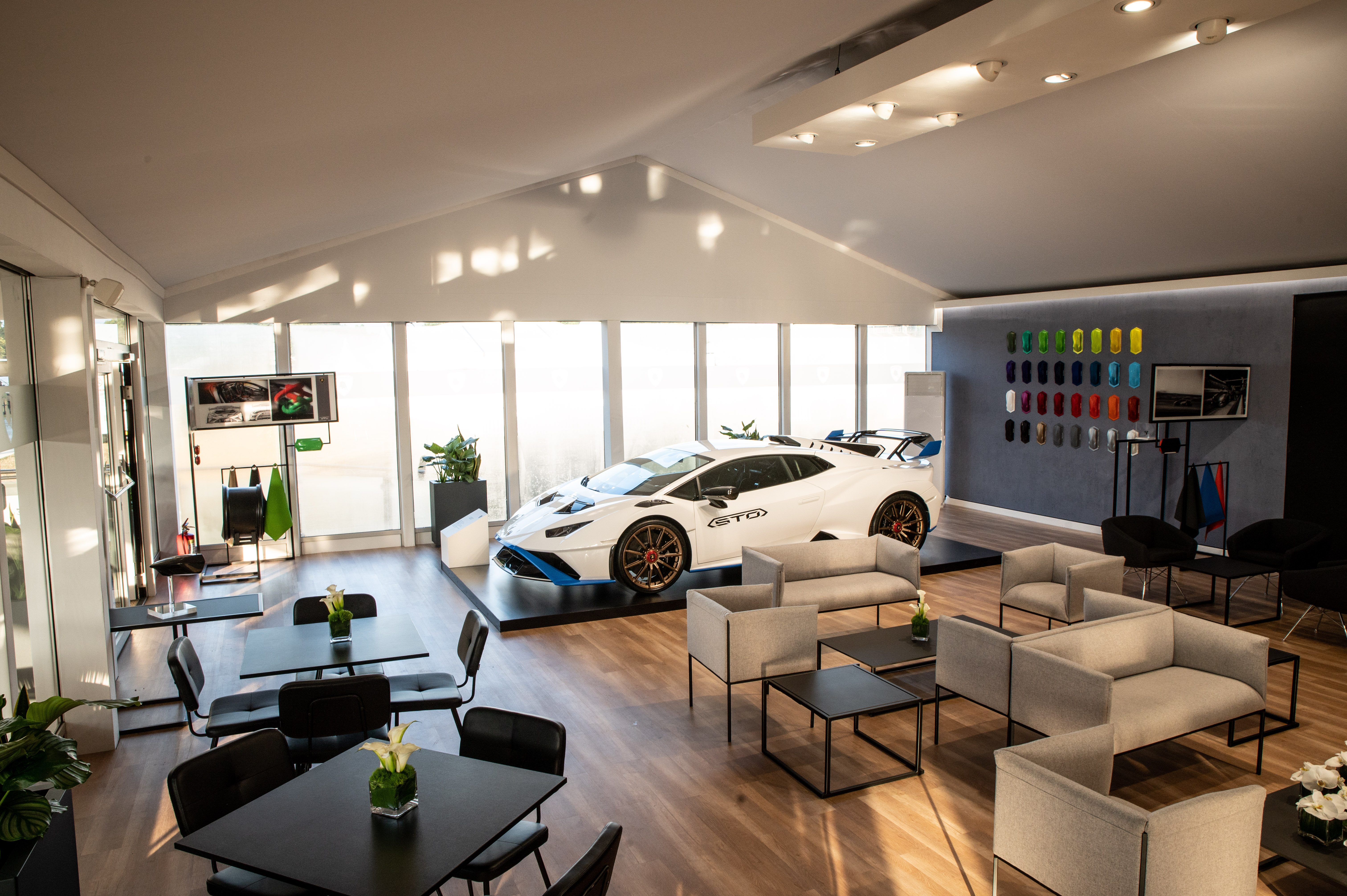 A white Lamborghini Huracán STO is displayed on a platform in a modern, spacious exhibition tent with lounge seating and large windows.