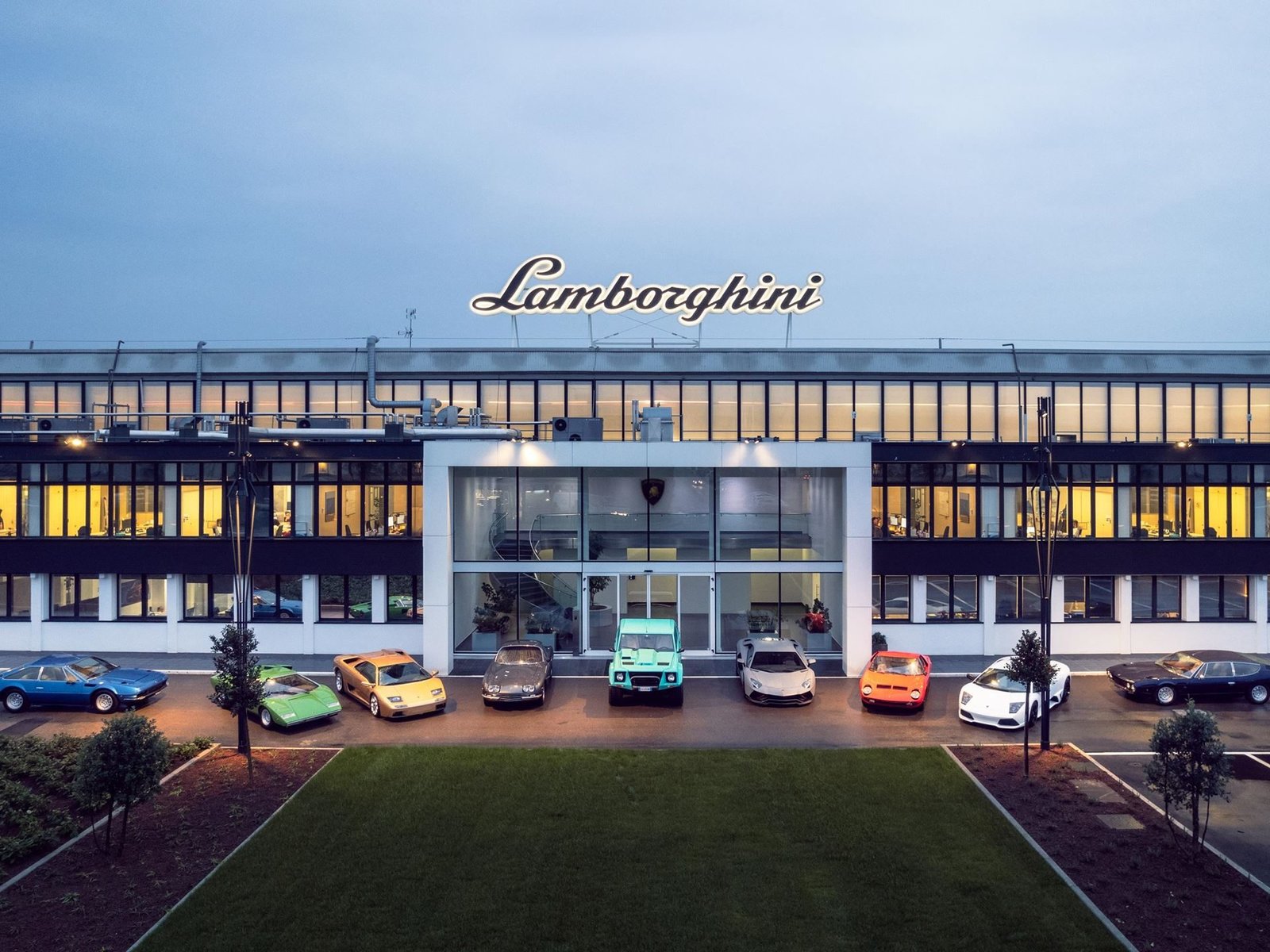 Classic and modern Lamborghini vehicles lined up in front of the illuminated Sant'Agata Bolognese factory at dusk