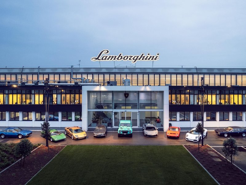 Classic and modern Lamborghini vehicles lined up in front of the illuminated Sant'Agata Bolognese factory at dusk