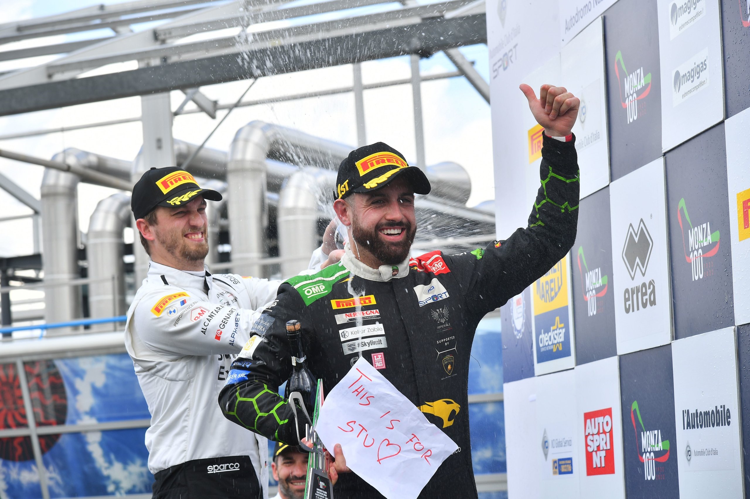 Di Folco'S Solo Monza Victory Proves The Huracán Gt3 Evo Still Bites Hard 3 Two racing drivers celebrate on the podium, one spraying champagne while holding a sign reading 'this is for stu' with a heart