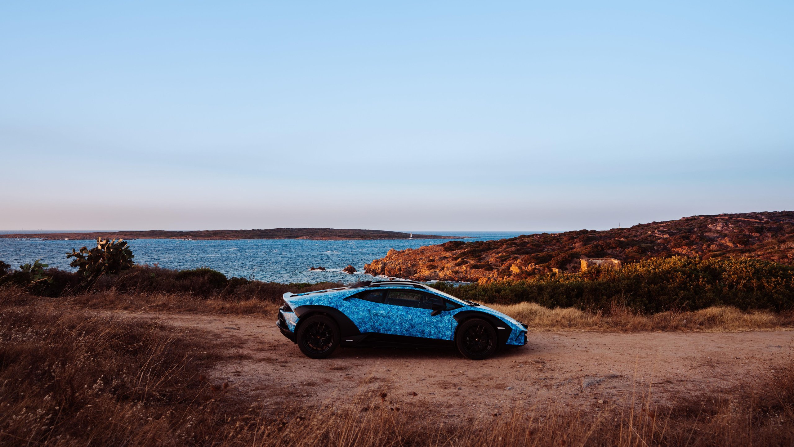 370 Hours Of Blue: How Lamborghini'S Opera Unica Sterrato Turns Paint Into Philosophy 9 Blue crystal-effect opera unica lamborghini huracán sterrato parked on a dirt path overlooking a calm sardinian sea under a clear sky