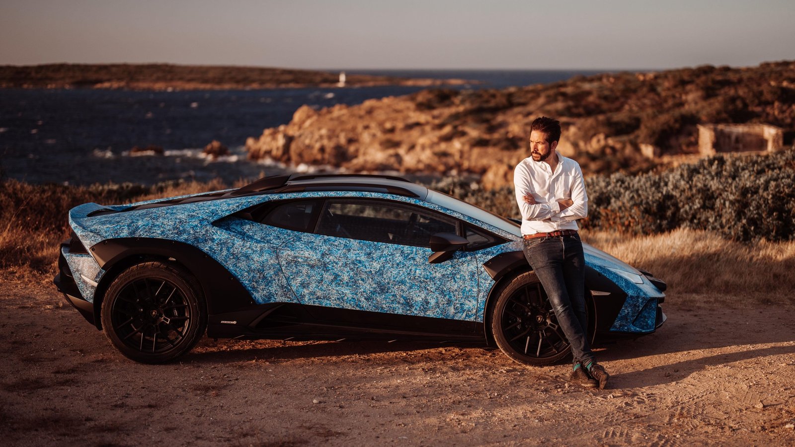 A man leans against the blue crystal-effect Opera Unica Lamborghini Huracán Sterrato on a Sardinian dirt path overlooking the sea at sunset