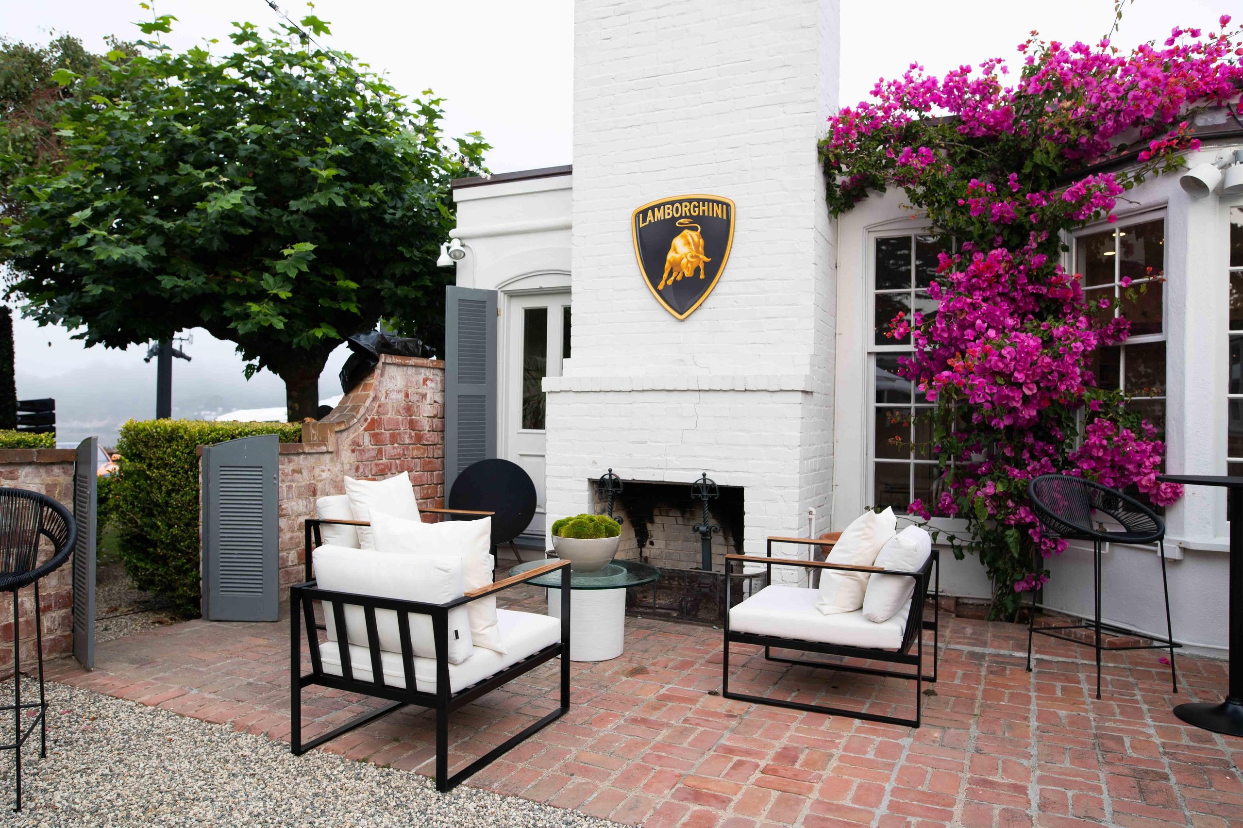 Lamborghini lounge monterey outdoor seating area with white chairs, lamborghini shield logo on white brick wall, and pink bougainvillea
