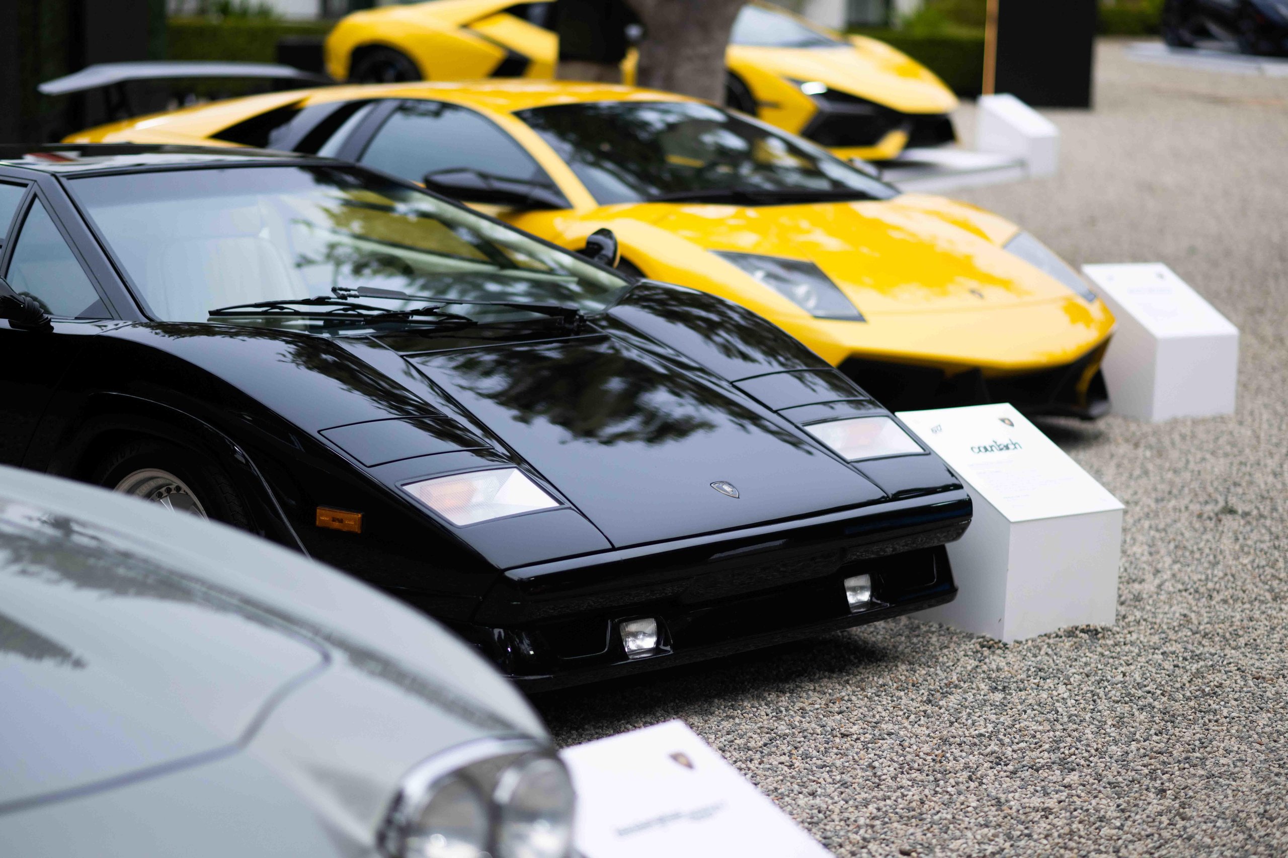 Black lamborghini countach in the foreground with yellow murciélago and yellow revuelto behind it on gravel at the lounge monterey event