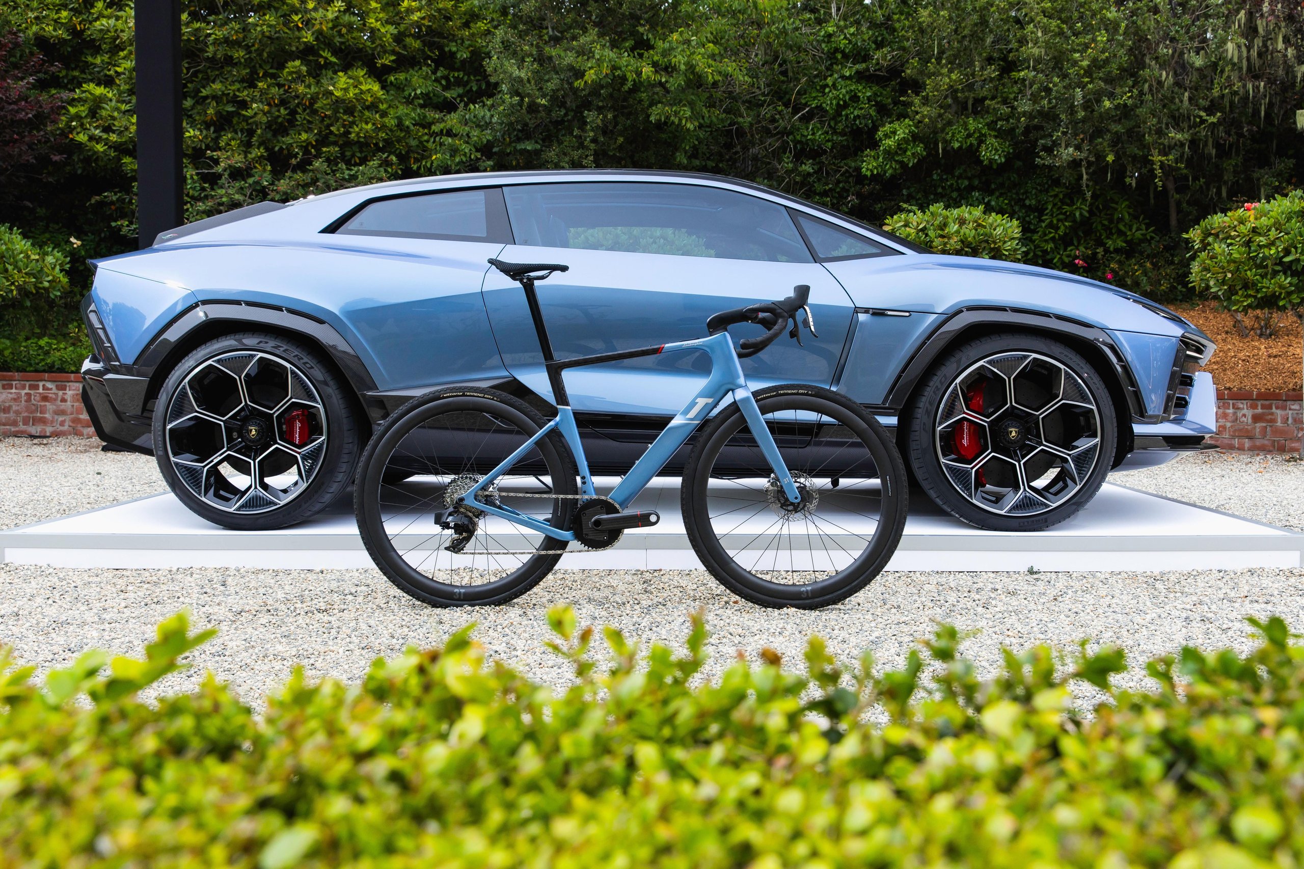Light blue lamborghini lanzador concept on white platform with matching 3t electric bicycle in the foreground against lush green background
