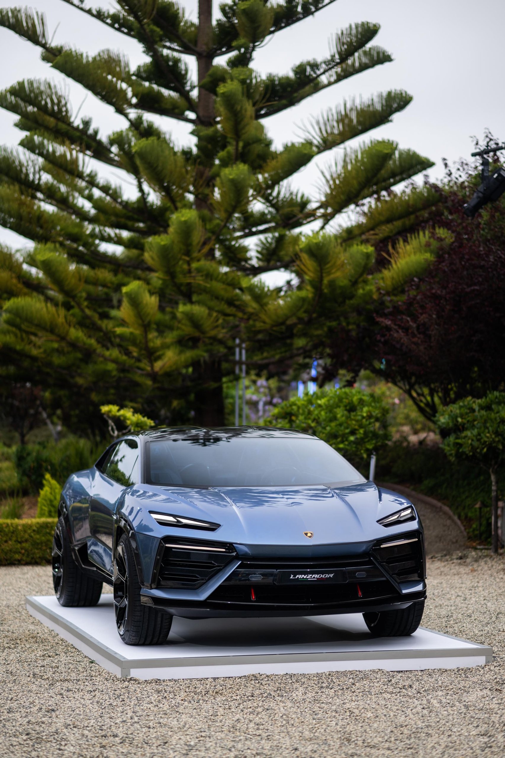 Front view of the lamborghini lanzador concept on a white display platform with lush green foliage in the background at monterey