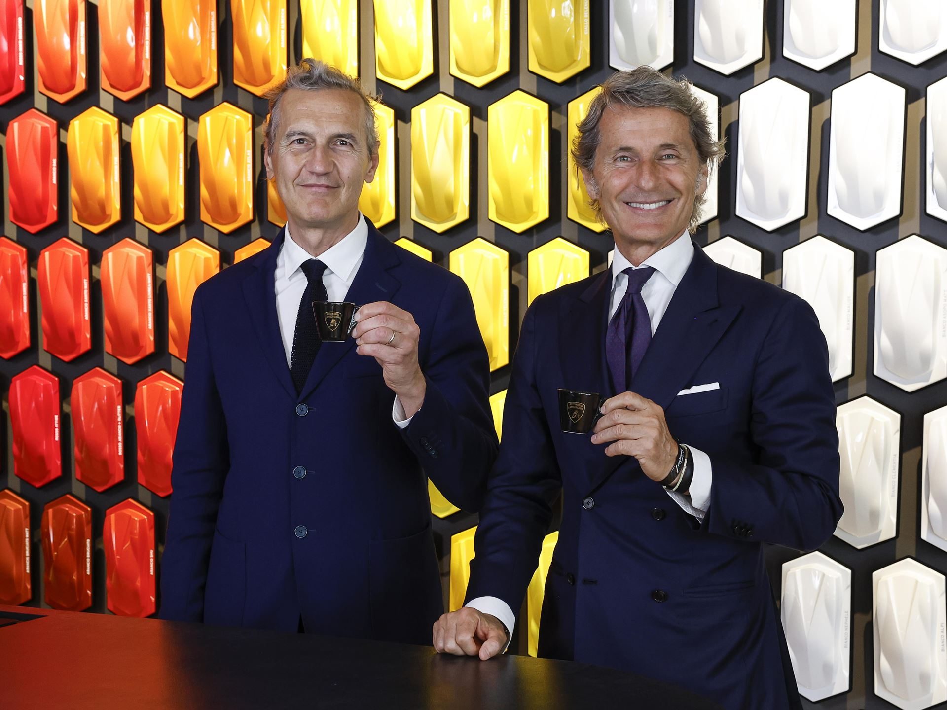 Lamborghini Partners With Lavazza To Pour Italian Luxury Beyond The Showroom 3 Two executives holding lamborghini-branded espresso cups in front of a colorful car paint sample display wall